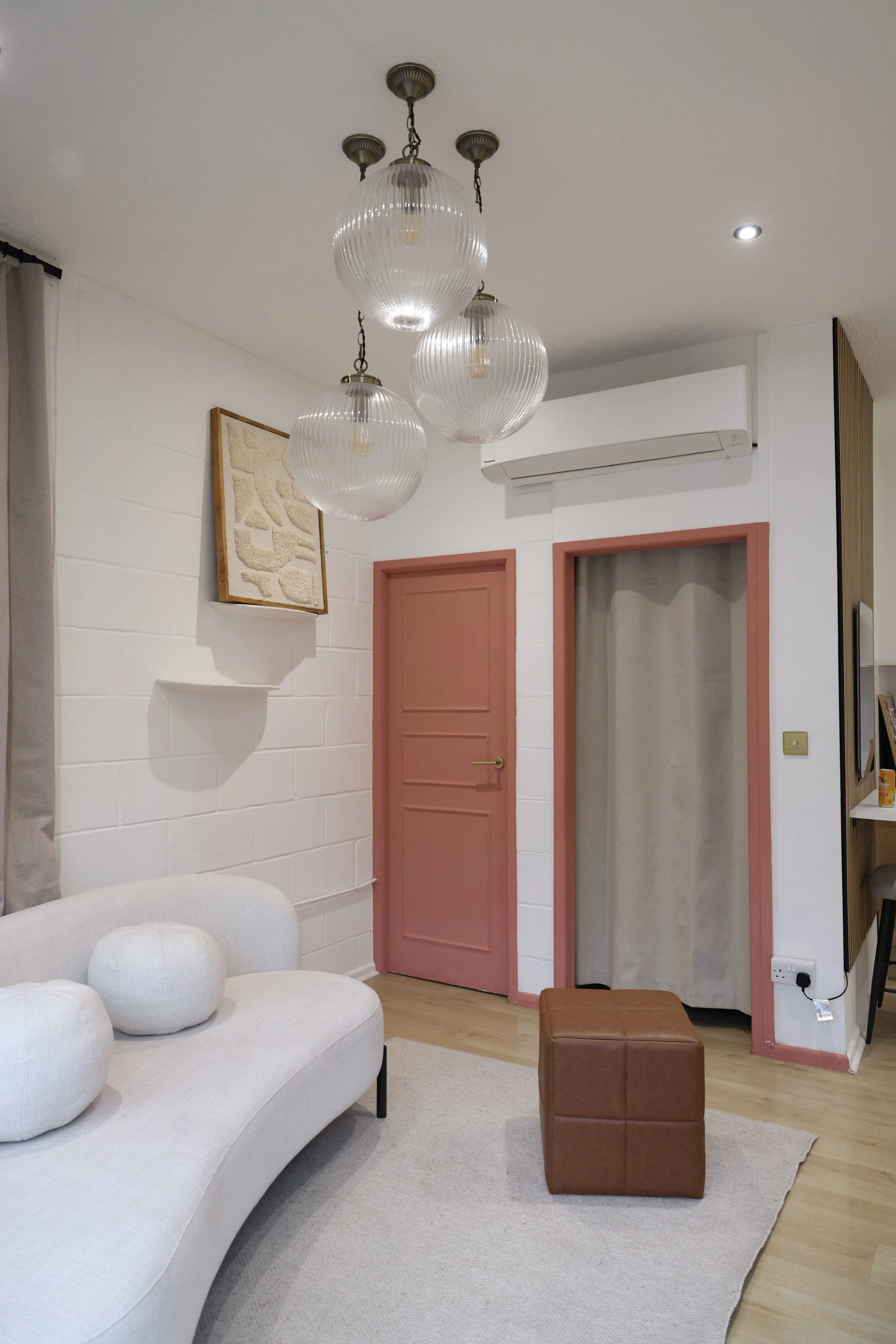 Living room with white brick wall, pink door and doorway, beige sofa with round pillows, brown ottoman, hanging glass globe lights, air conditioning unit, framed artwork, beige curtains, and wooden floor.