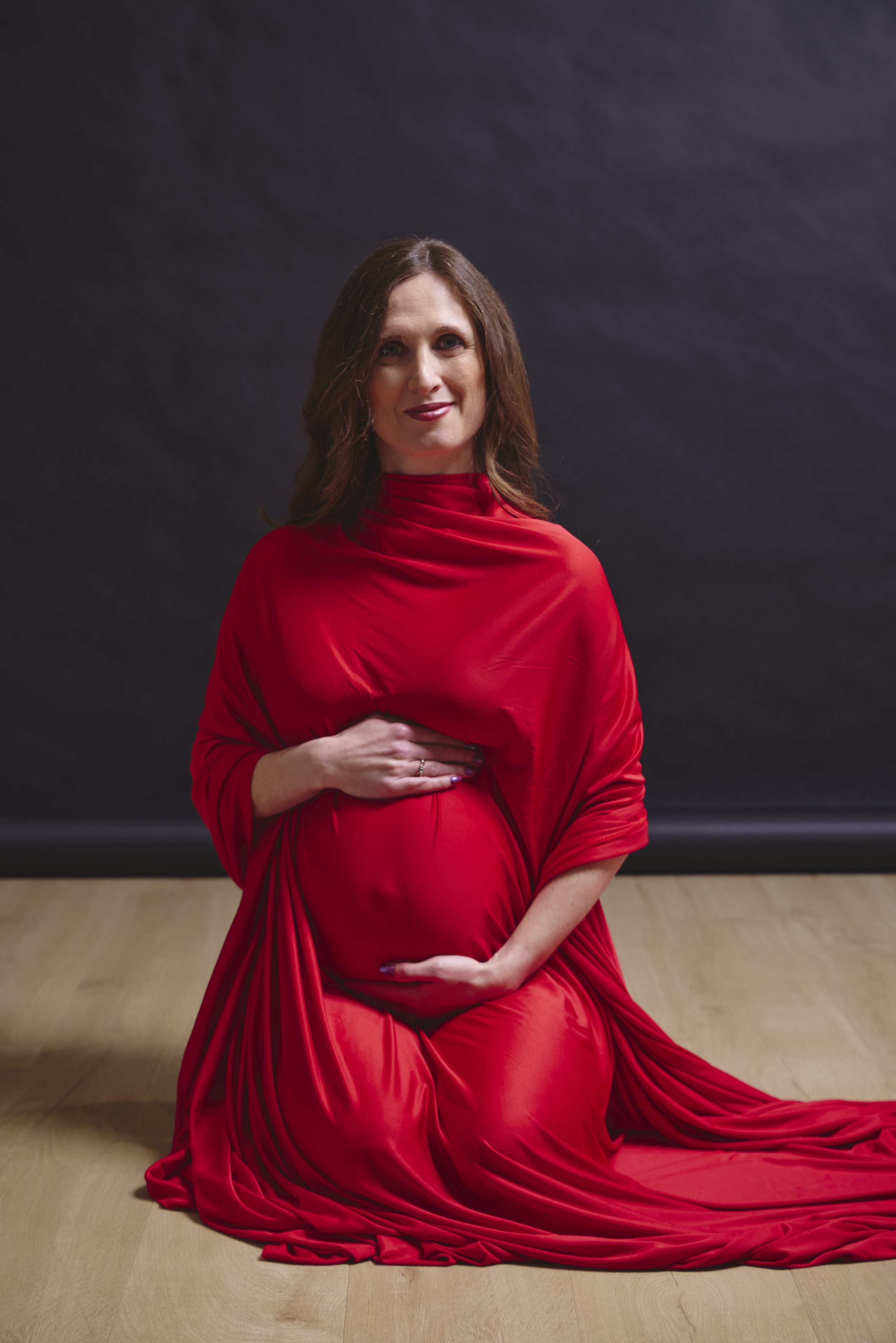 Pregnant woman in a red dress sitting on the floor against a dark background.