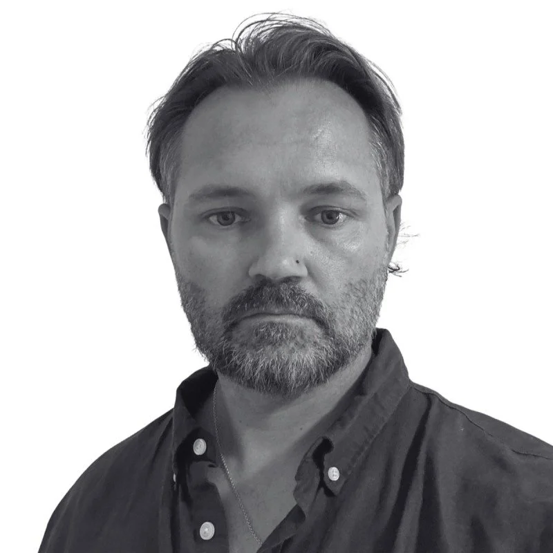 A black and white portrait of a Jarrod Guyer with medium-length hair, a beard, wearing a collared shirt, looking directly at the camera with a serious expression.