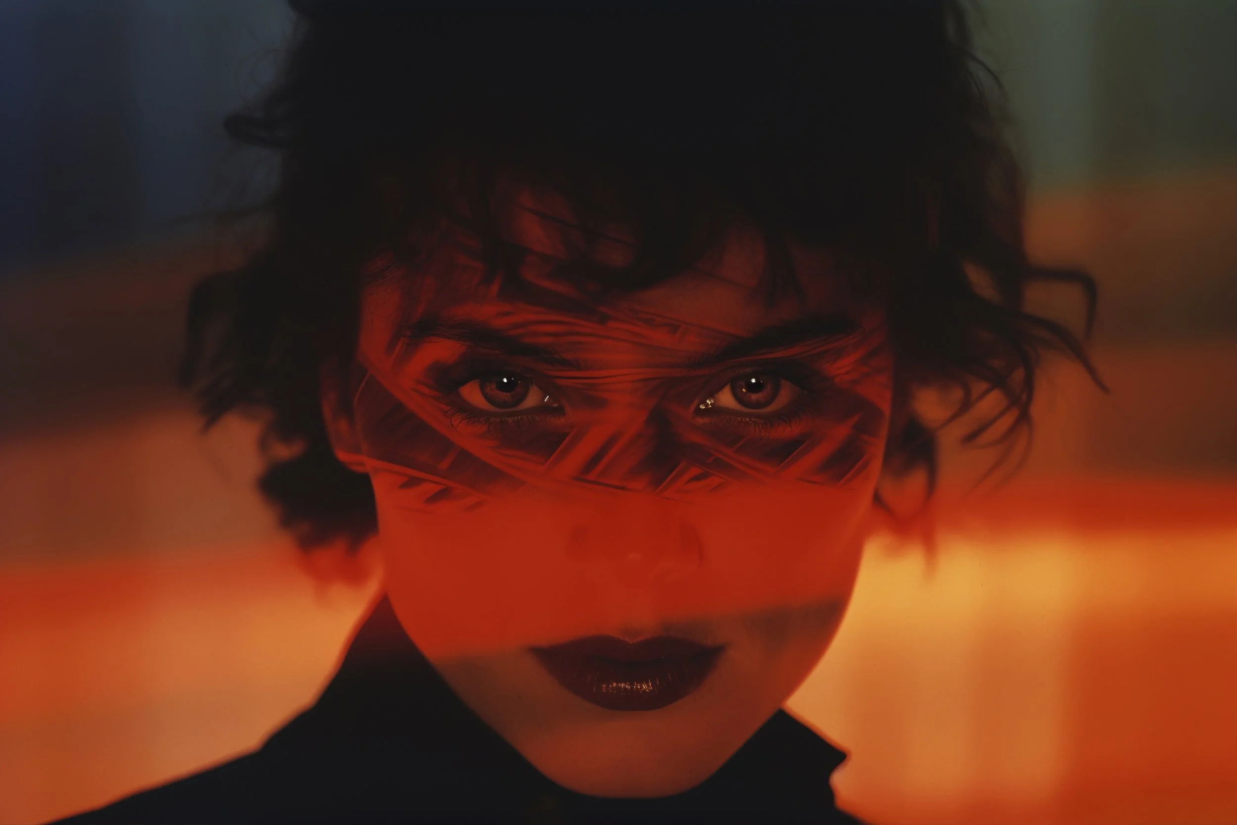 A woman with dark hair and dark lipstick wearing an orange face covering with black geometric patterns, looking directly at the camera against a vibrant orange and reddish background.