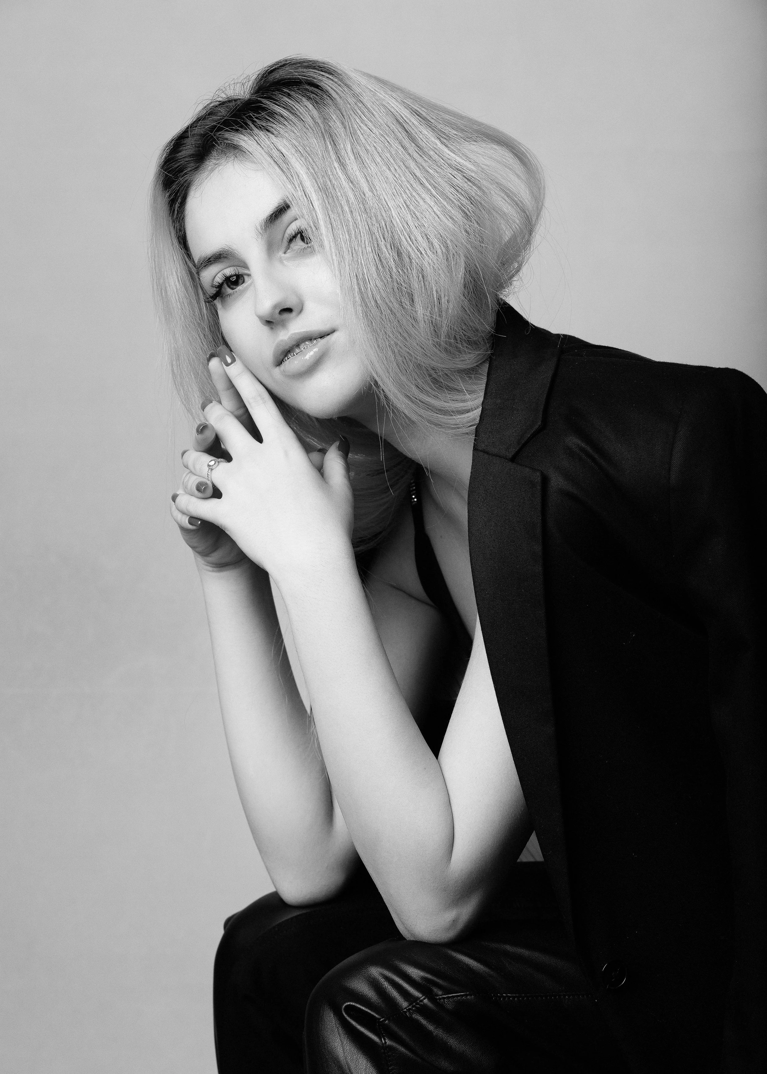 Black and white photo of a woman with shoulder-length hair, wearing a blazer and leather pants, posing with her hand near her face.