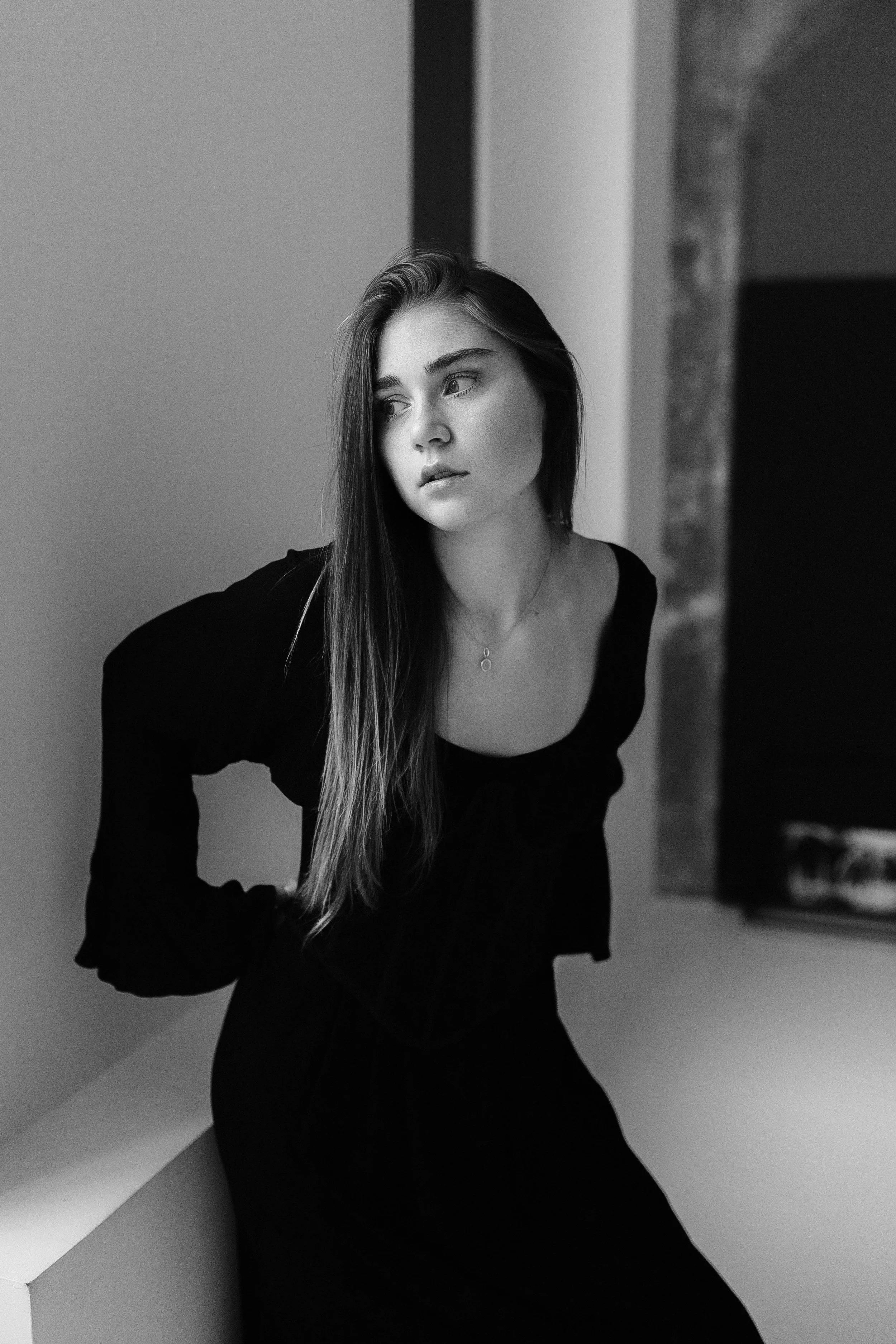 Black and white photograph of a young woman with long hair, wearing a black top, sitting indoors near a wall with her body slightly turned and looking away from the camera.