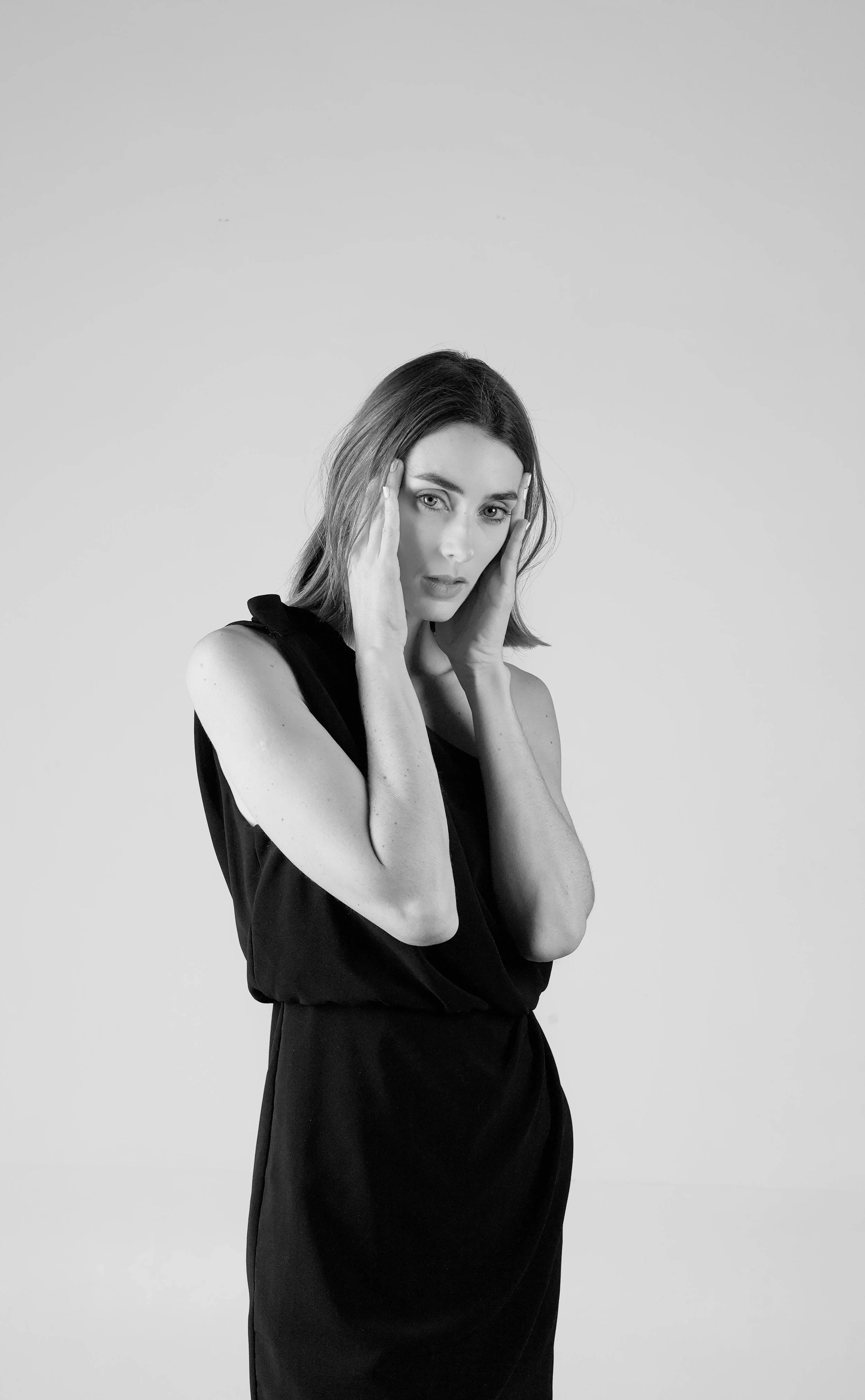 A woman in a black sleeveless top and skirt holding her head with both hands, looking at the camera, in a black-and-white studio portrait.