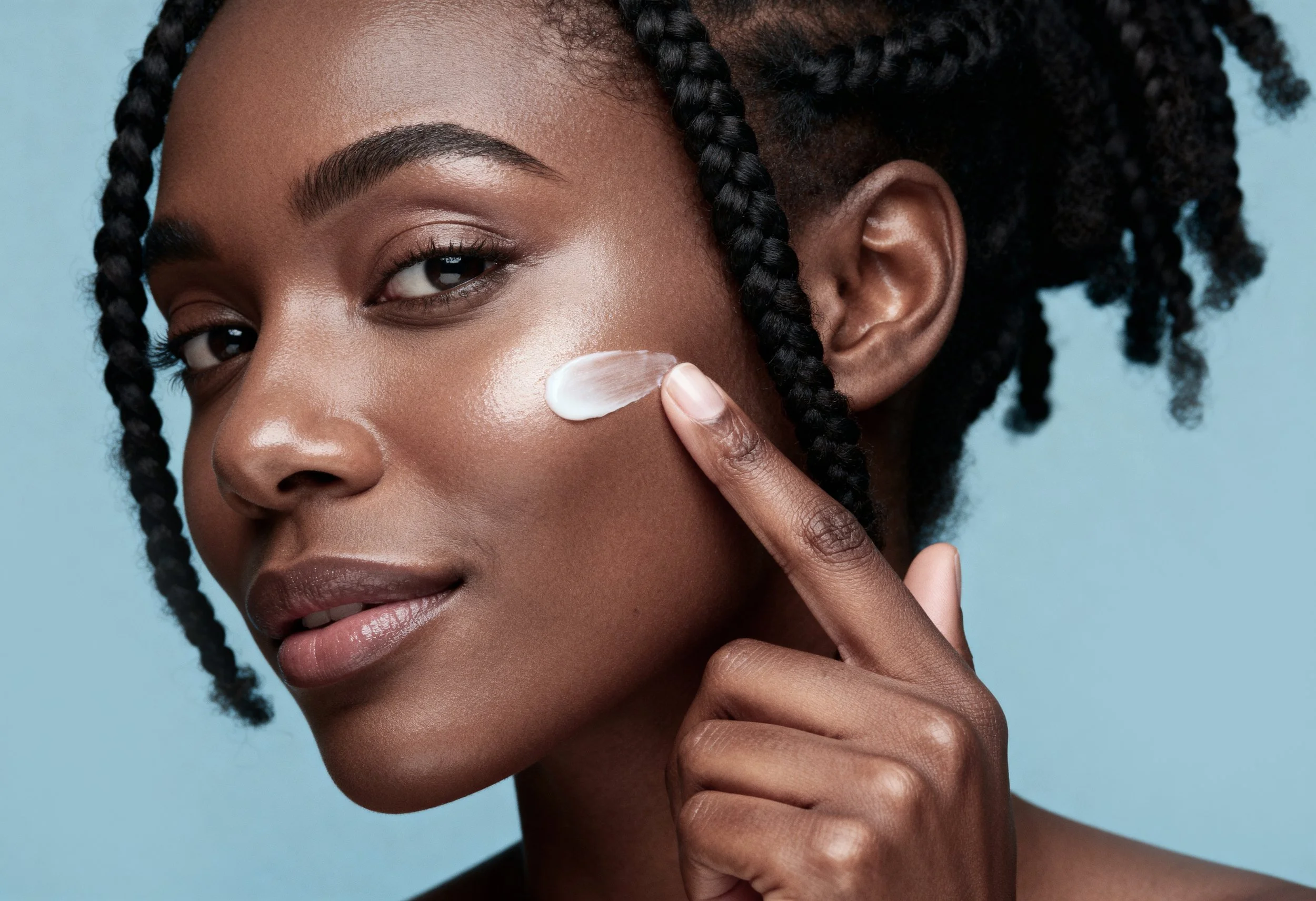A woman with dark skin and braided hair applying lotion to her face against a light blue background.