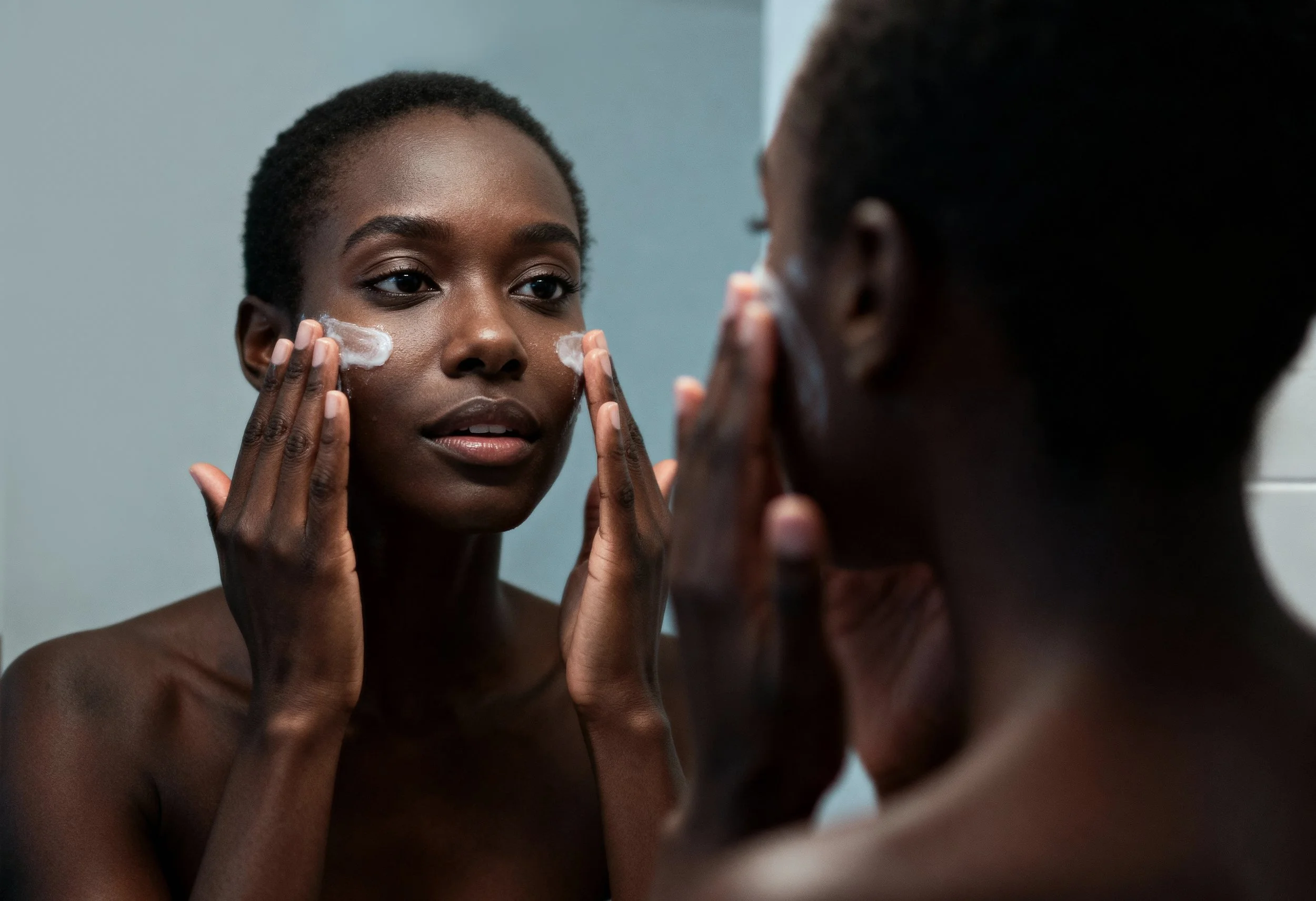 A young woman with short hair applying facial cleanser in front of a mirror, with her reflection visible.