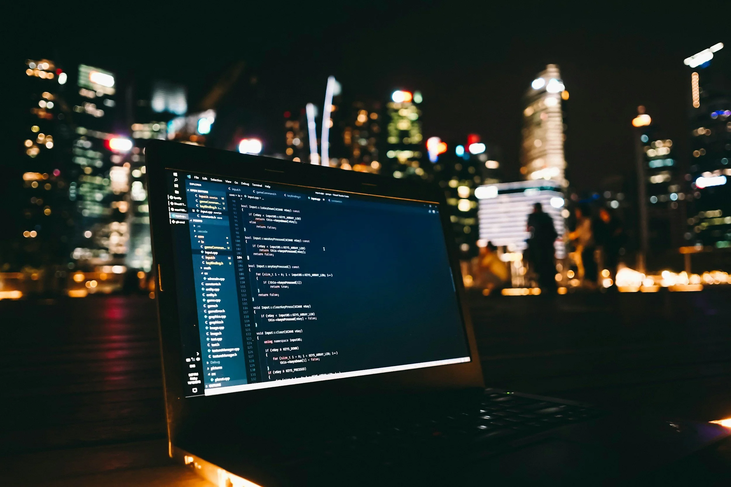 Laptop with code on screen in front of a city skyline at night, with out-of-focus buildings and city lights in the background.