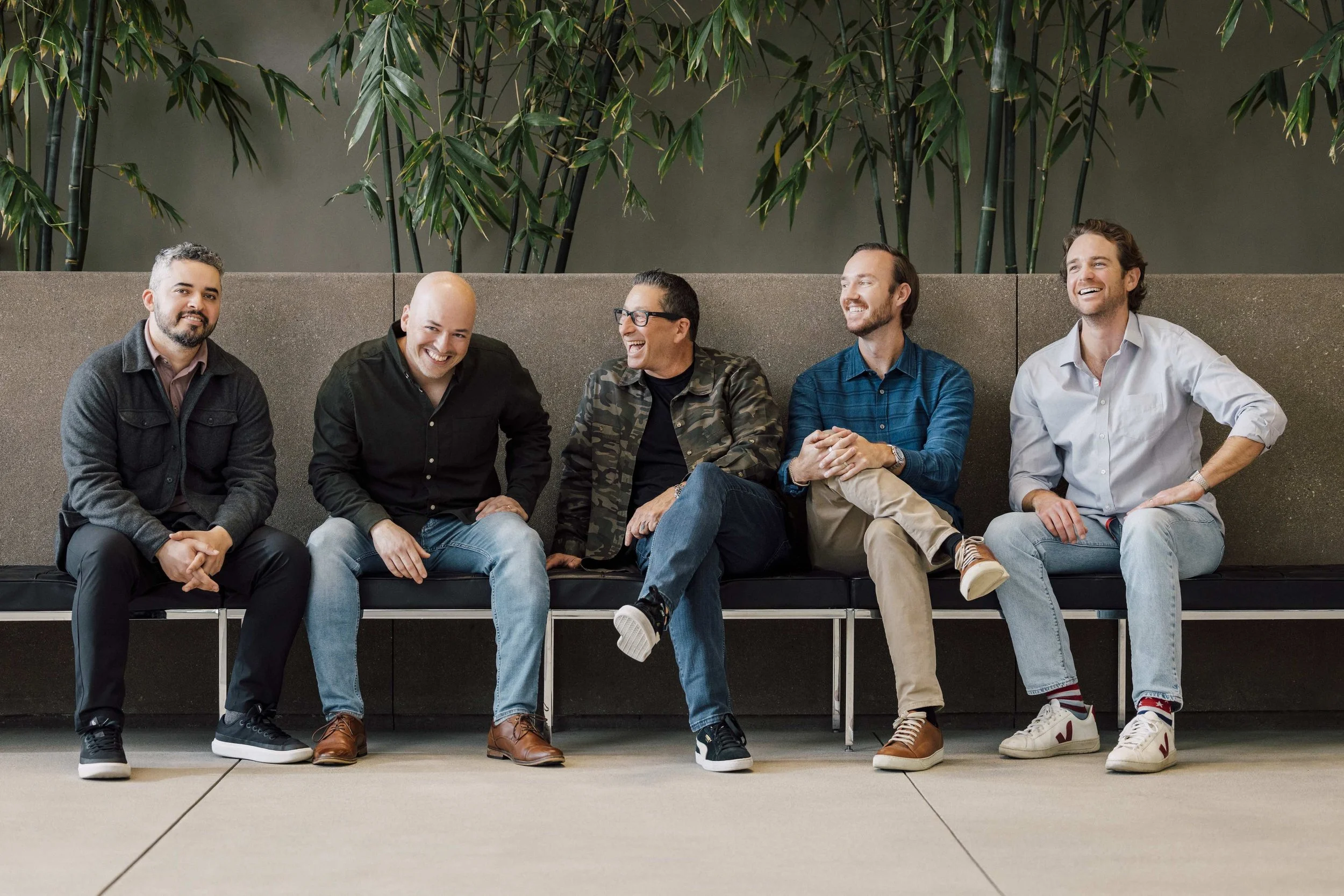 Five men sitting on a bench, smiling and laughing, in a modern indoor setting with green plants in the background.