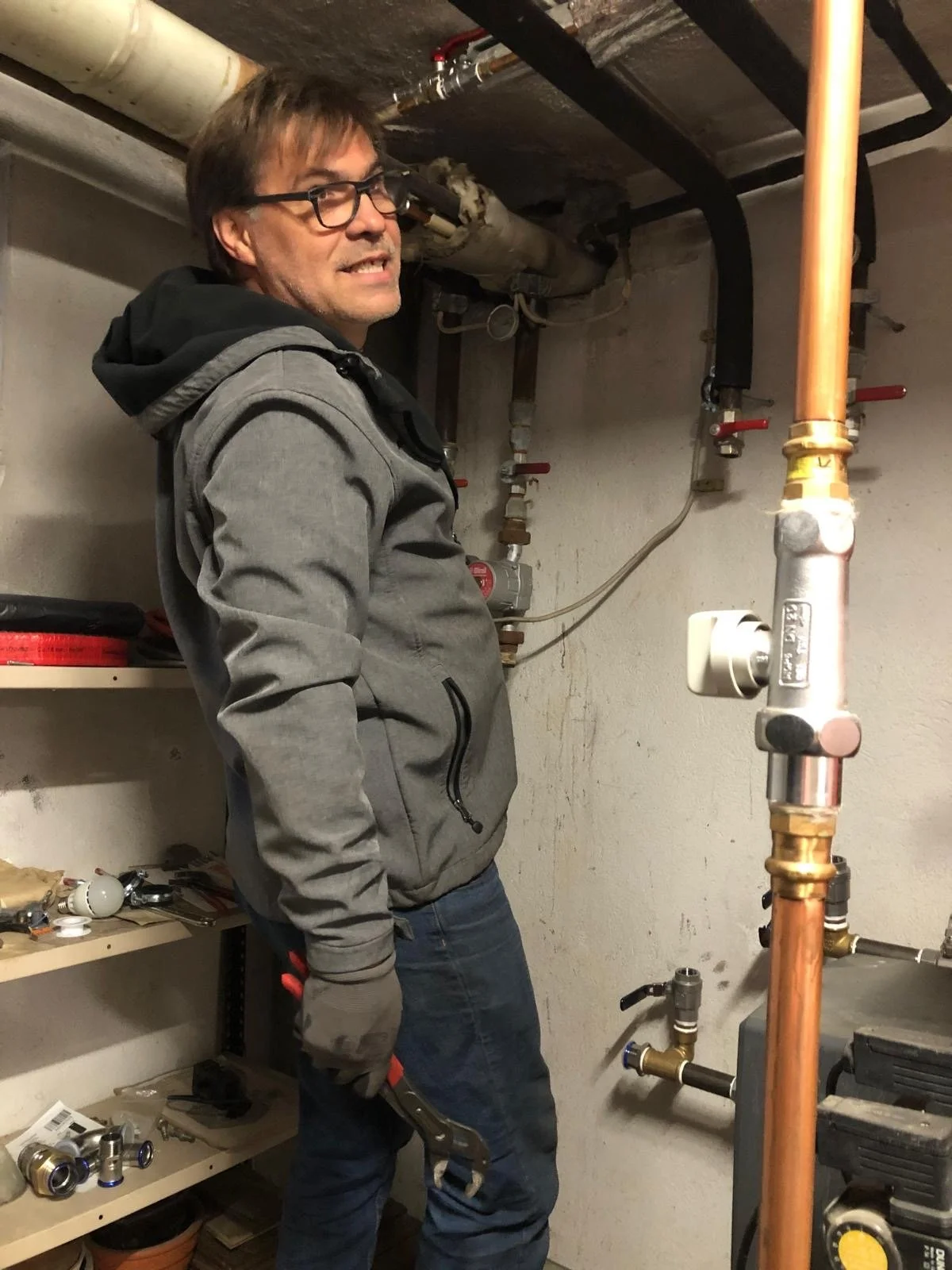 Mann in dunkler Brille und grauer Jacke arbeitet an Wasserrohrleitungen in einem Kellerraum.