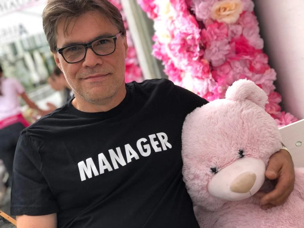 Mann trägt ein schwarzes T-Shirt mit der Aufschrift "MANAGER" und hält ein pinkes Plüschtier, im Hintergrund sind pinke Blumen