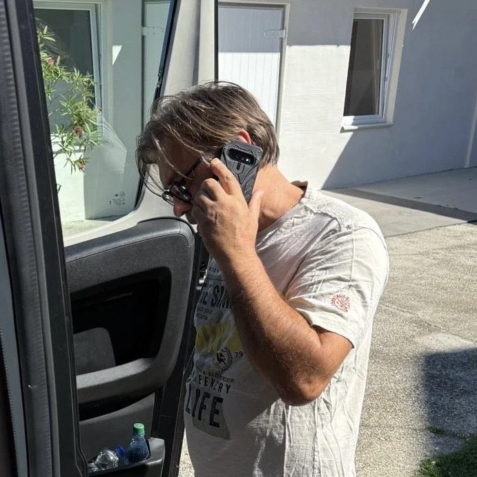 Ein Mann steht neben einem Auto, spricht mit einem Handy am Ohr und trägt eine Sonnenbrille.