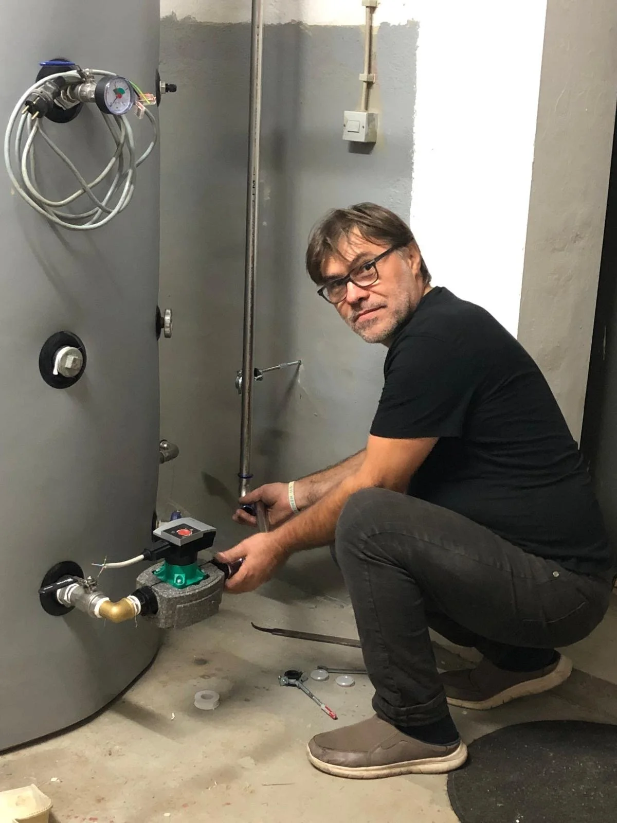 Ein Mann mit Brille und grauem Bart, der an einem Heizkessel arbeitet, während er in die Kamera schaut.