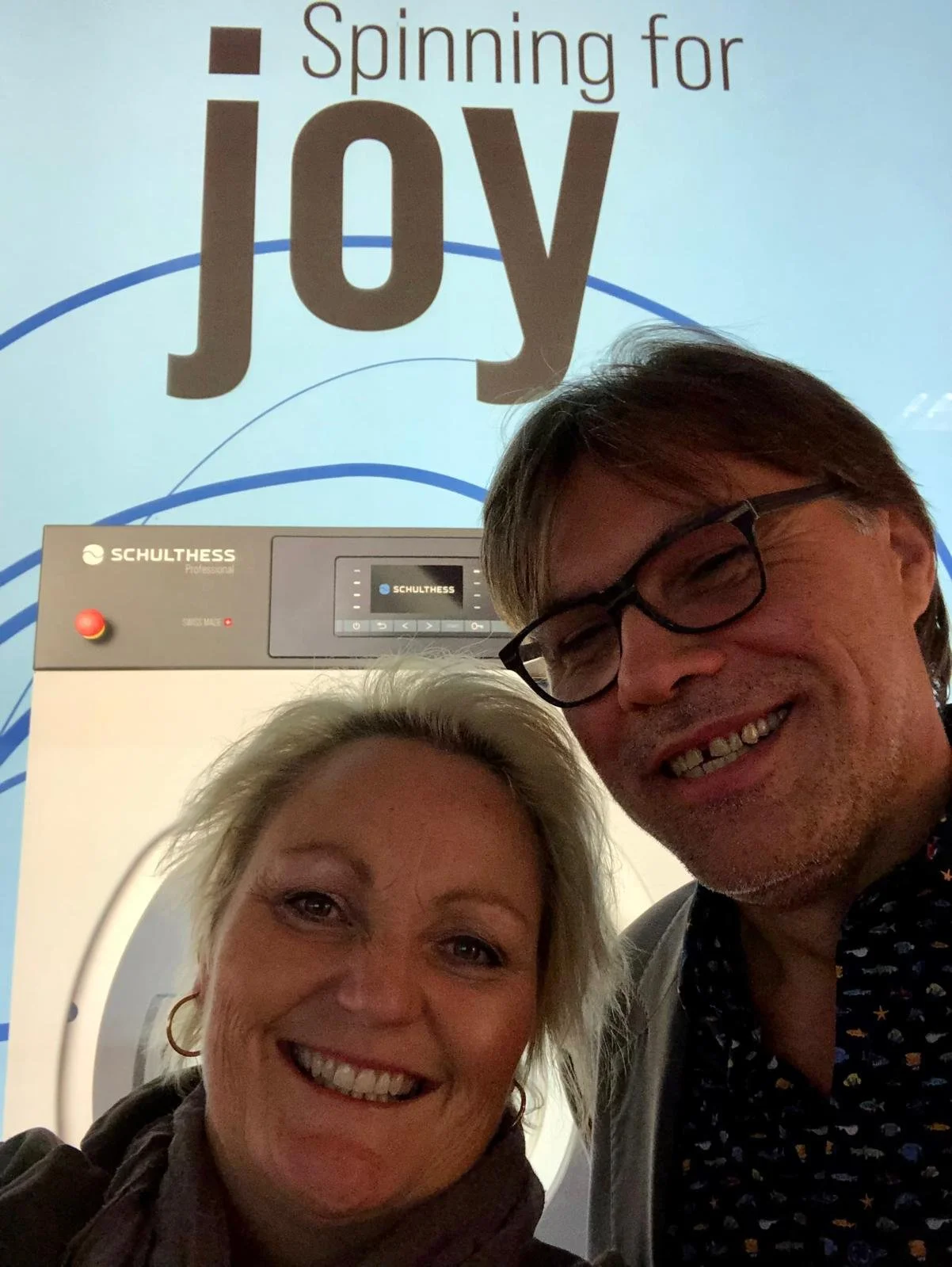 Zwei Personen, ein Mann und eine Frau, lächeln vor einem Hintergrund mit der Aufschrift "Spinning for Joy" und einem Bild eines Waschmaschinentronic. Der Mann trägt Brille und hat braunes Haar. Die Frau hat blondes Haar und trägt eine dunkle Jacke und Ohrringe.