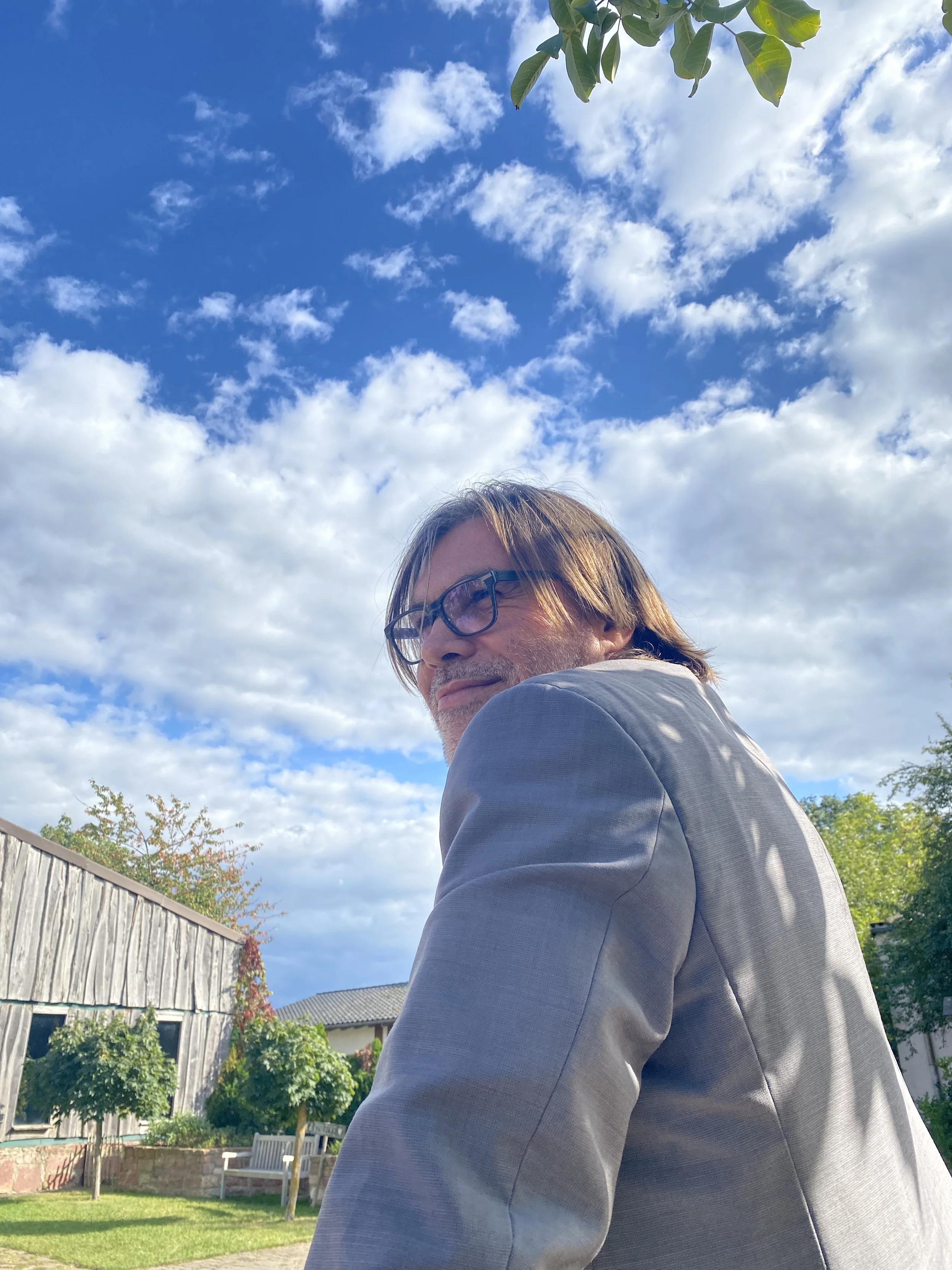 Ein Mann mit Brille trägt einen grauen Anzug und sitzt im Freien, im Hintergrund sind Bäume, ein Rasen und ein Gebäude, während der Himmel mit Wolken bedeckt ist.