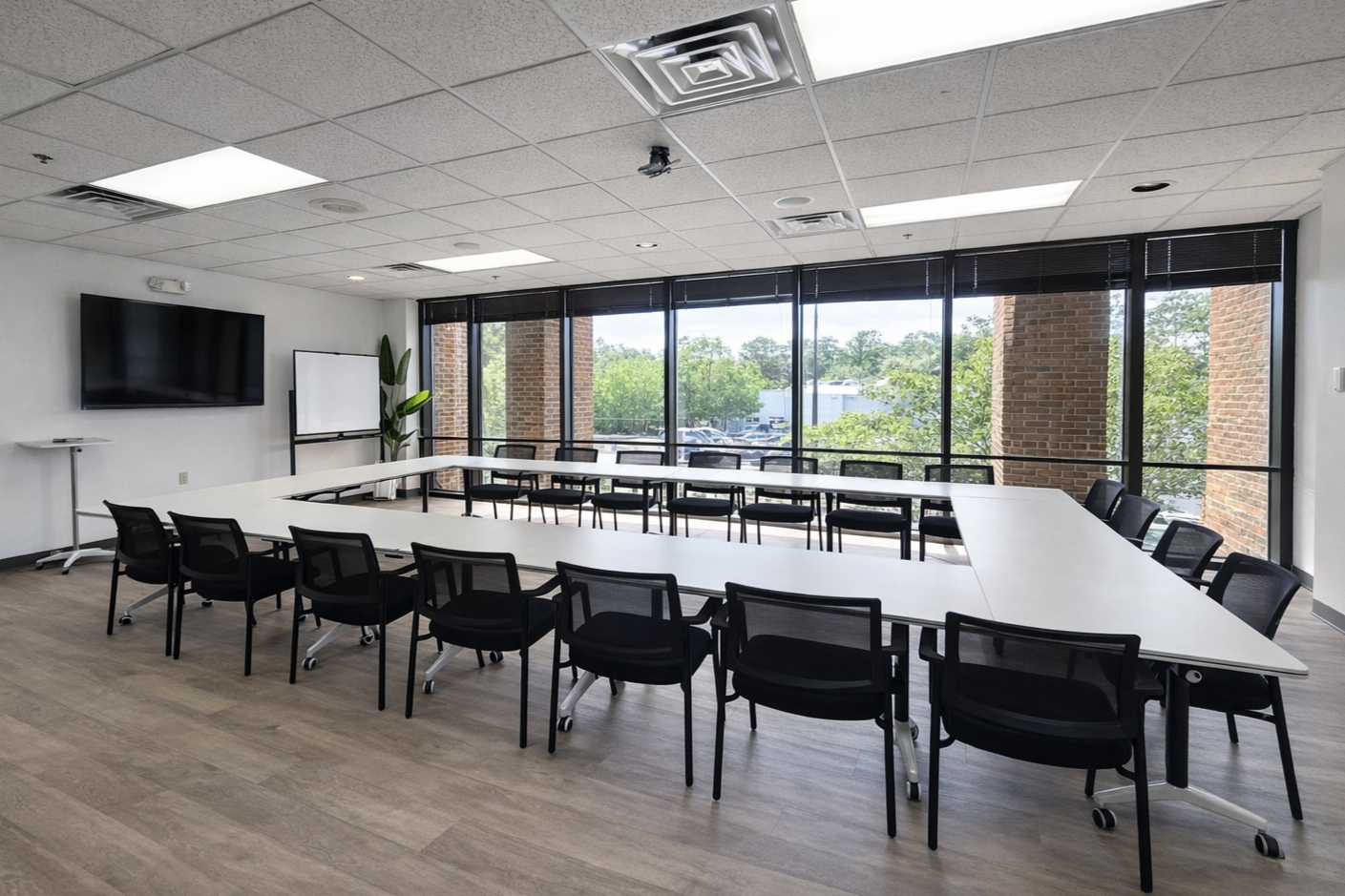 Empty modern conference room with U-shaped table, black chairs, large windows, wall-mounted TV, whiteboard, and a potted plant.