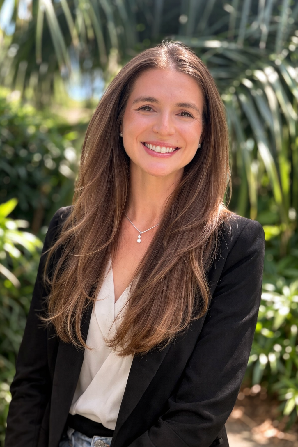 Portrait of a smiling young woman with long brown hair, wearing a black blazer, white blouse, and a silver necklace with a small pendant, standing outdoors with green foliage in the background.