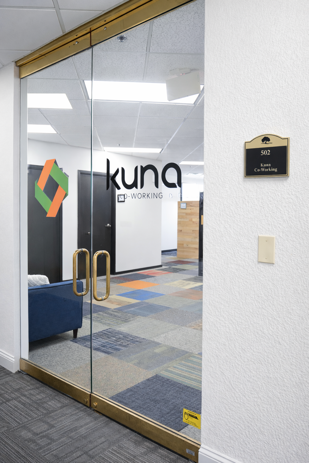 Glass door with a company logo and text that says 'Kuna Co-Working', leading into a modern office space with colorful carpet tiles, a blue sofa, and a wooden reception desk.