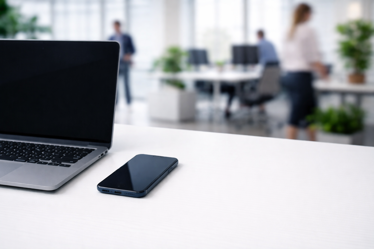 Imagen de un escritorio en una oficina moderna con una laptop y un teléfono móvil en primer plano, y personas trabajando en el fondo.