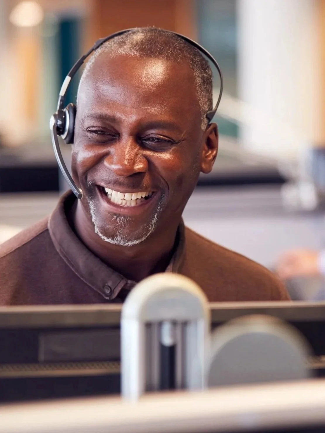 An older man with a smile, wearing a headset, appears to be working at a computer in an office setting.
