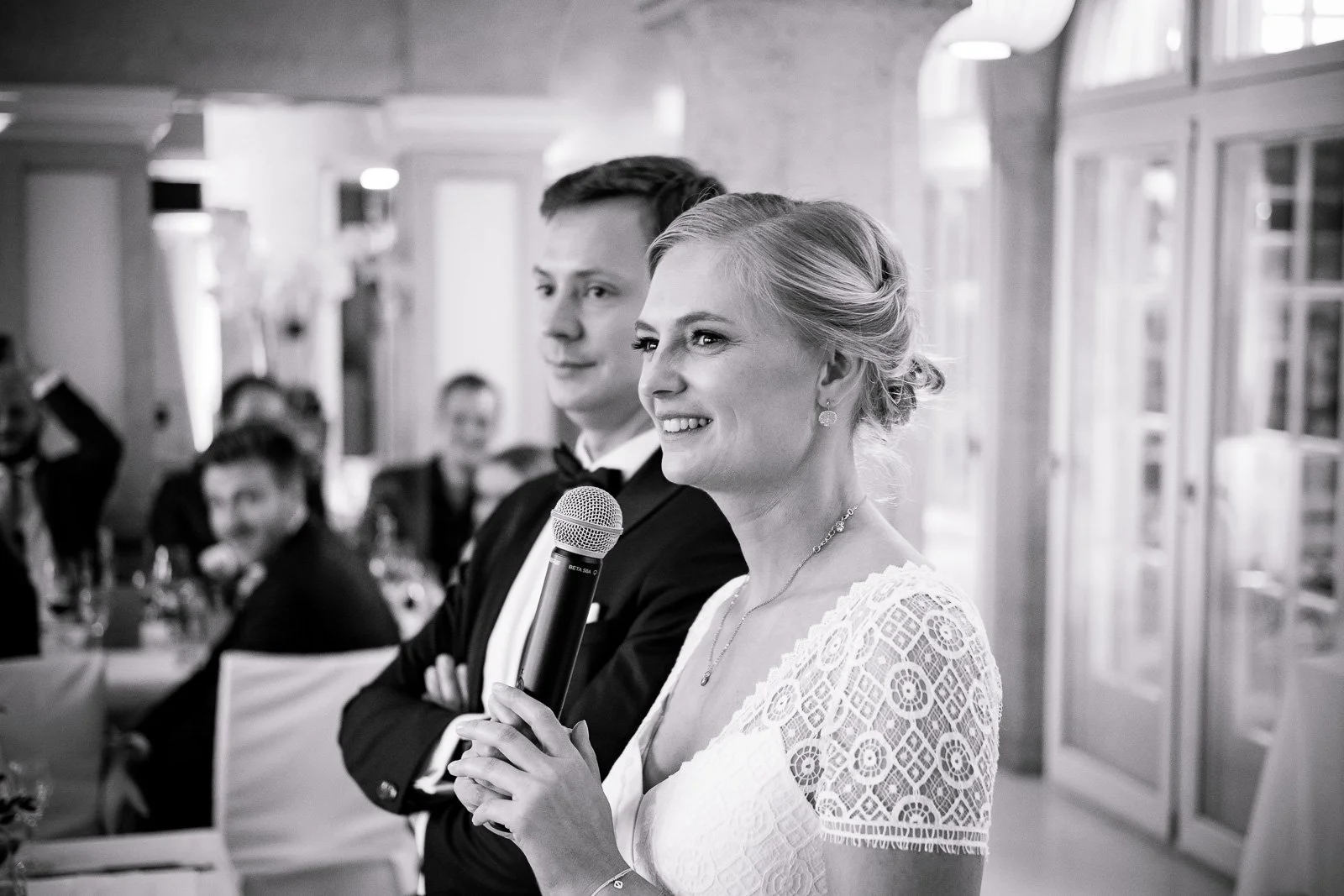 Braut hält Mikrofon bei Hochzeit, umgeben von Gästen in einem festlichen Raum.