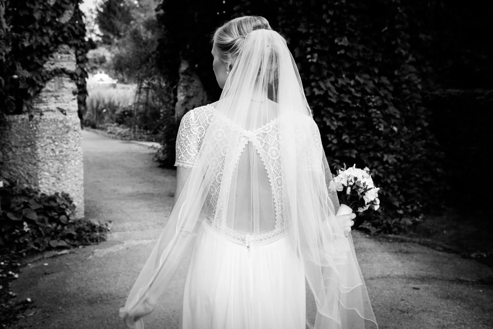 Junge Frau in einem weißen Hochzeitskleid mit Schleier, die eine Hand nach hinten hält und in der anderen ein Blumenbouquet trägt, beim Spaziergang auf einem Weg durch einen Garten oder Park.