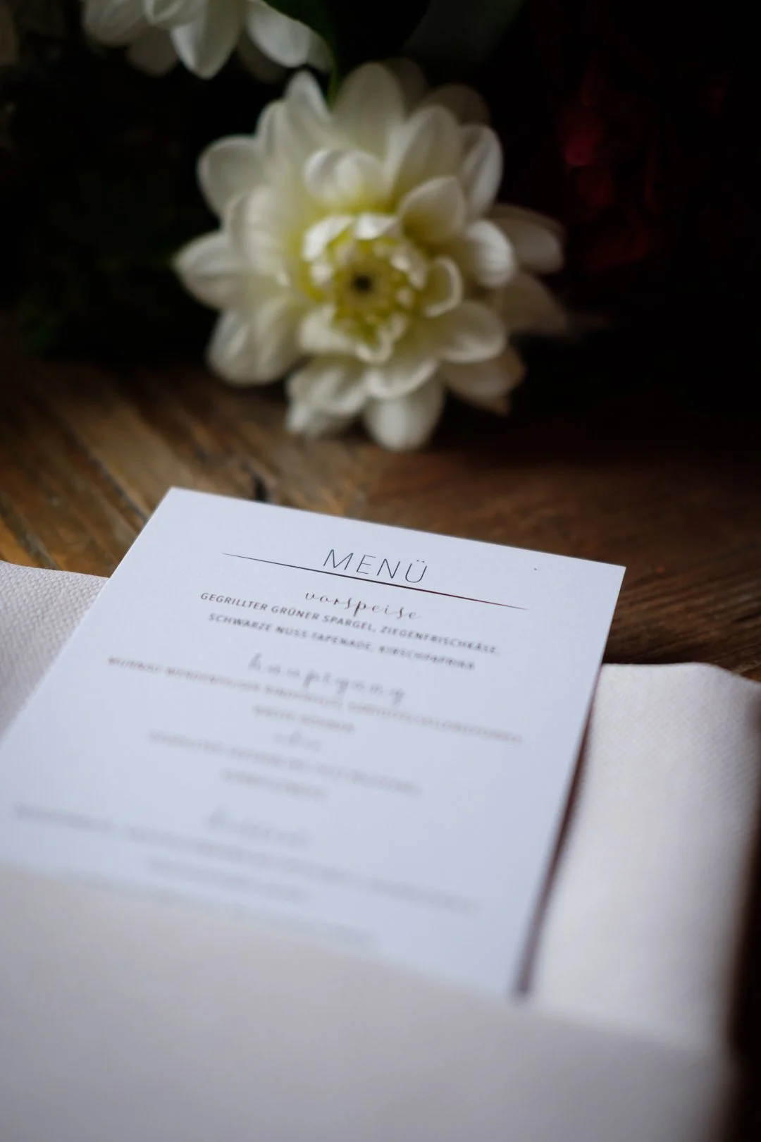 Ein Speisekartenbogen liegt auf einer gedeckten Tafel, im Hintergrund eine weiße Blume, wahrscheinlich eine Dahlie.
