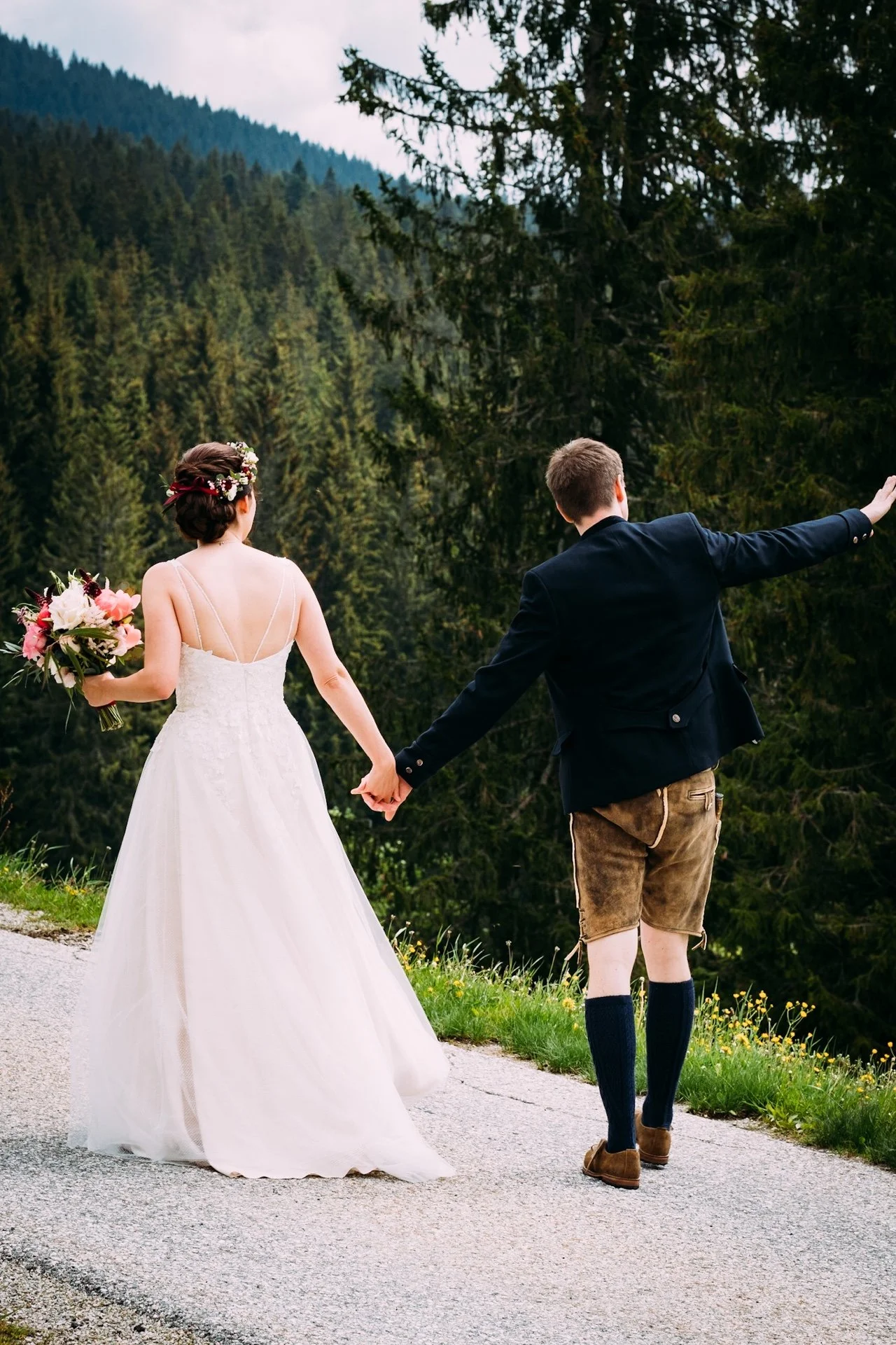 Ein Brautpaar hält Händchen, während es an einem Waldrand steht. Die Braut trägt ein weißes Hochzeitskleid und eine Blumenkrone, die sie mit einem Blumenstrauß in der Hand hält. Der Bräutigam trägt eine dunkle Jacke, eine Trachtenhose und kniehohe So