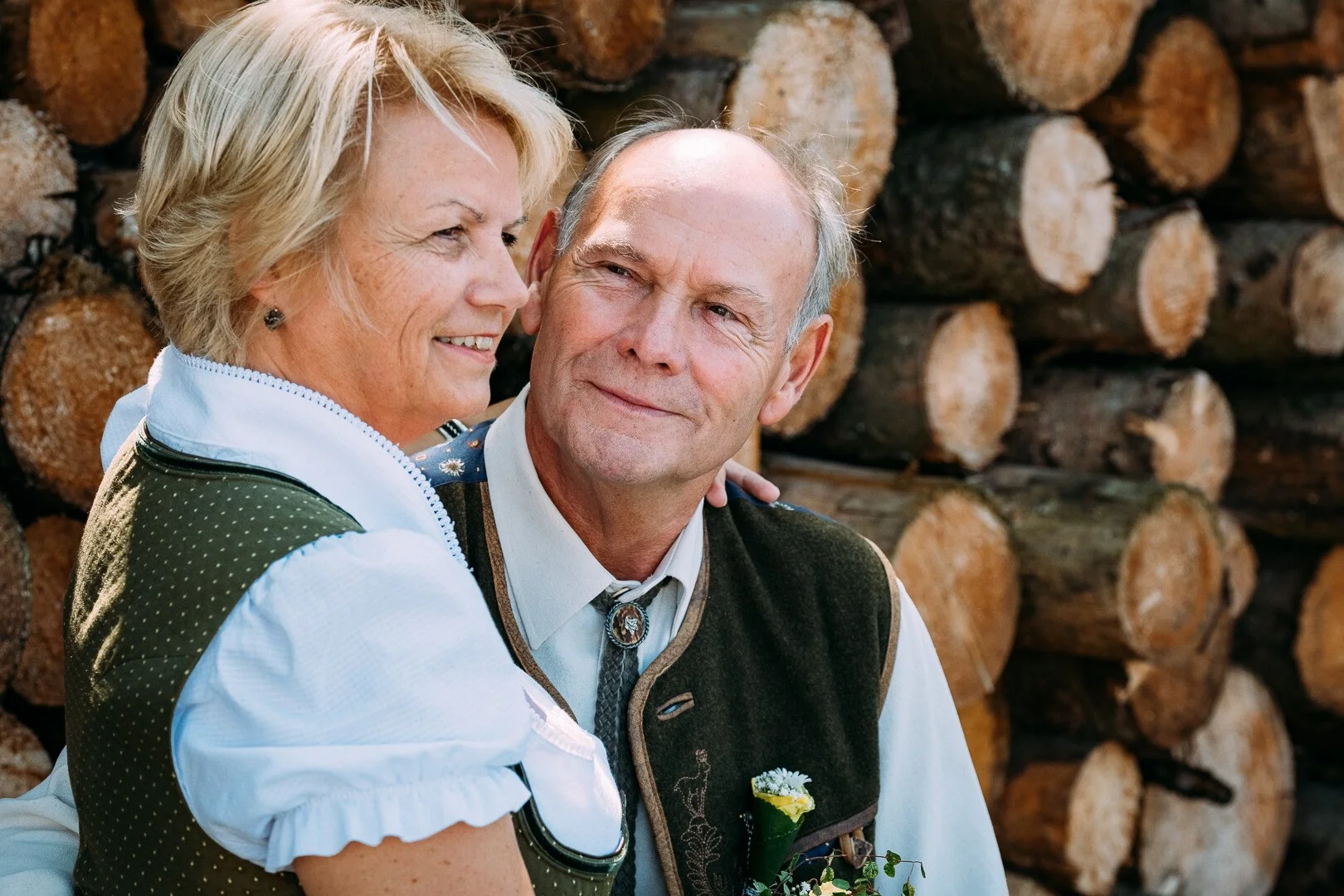 Ein älteres Paar trägt Dirndl Tracht und Lederhose und steht vor einem Stapel Holz. Sie lächeln sich liebevoll an.