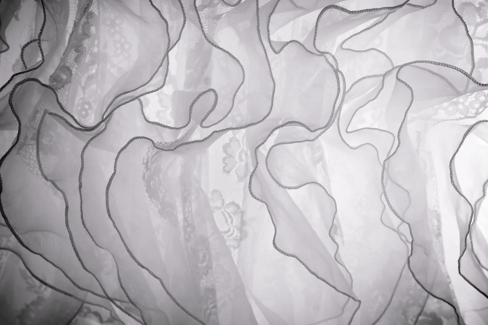 Feines weißes Tüllgewebe mit schwarzen Ziernähten, teils transparent, mit Wellenmustern.