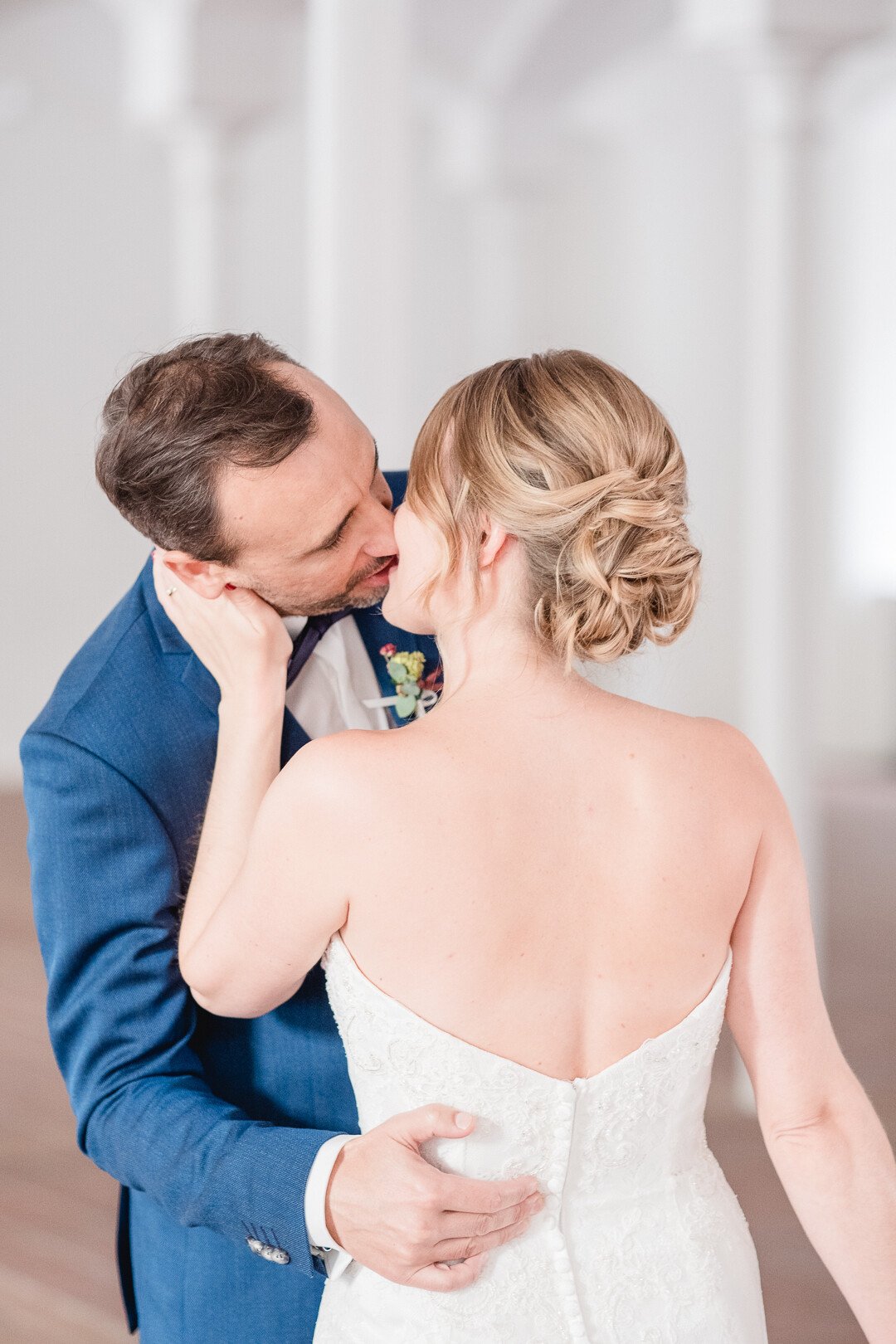 Ein Paar küsst sich bei einer Hochzeit, der Mann trägt einen blauen Anzug, die Frau ein weißes Kleid, sie sind umarmt.