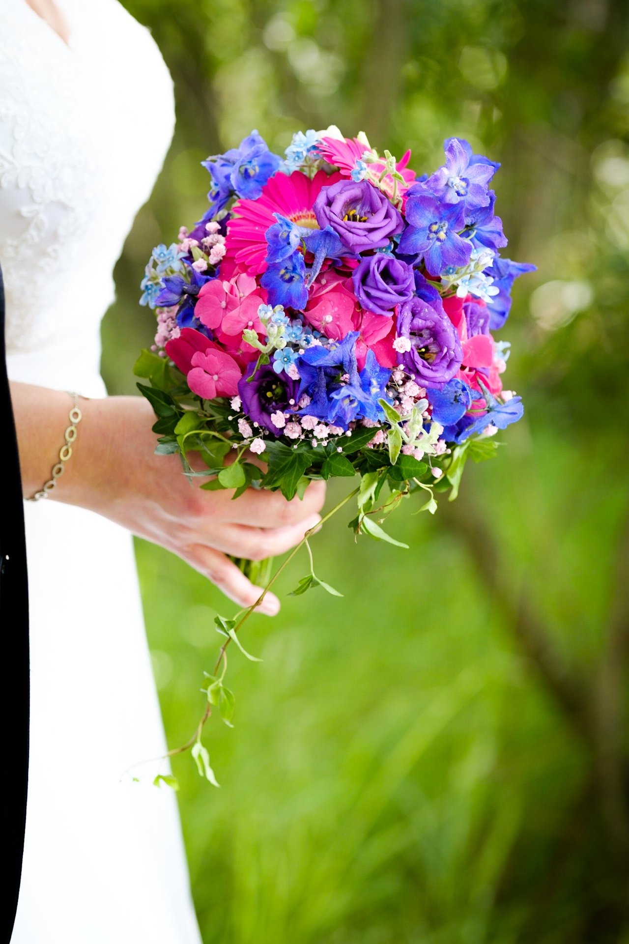 Eine Hand hält einen bunten Blumenstrauß mit lila, pinken und blauen Blumen vor einem grünen, unscharfen Hintergrund.