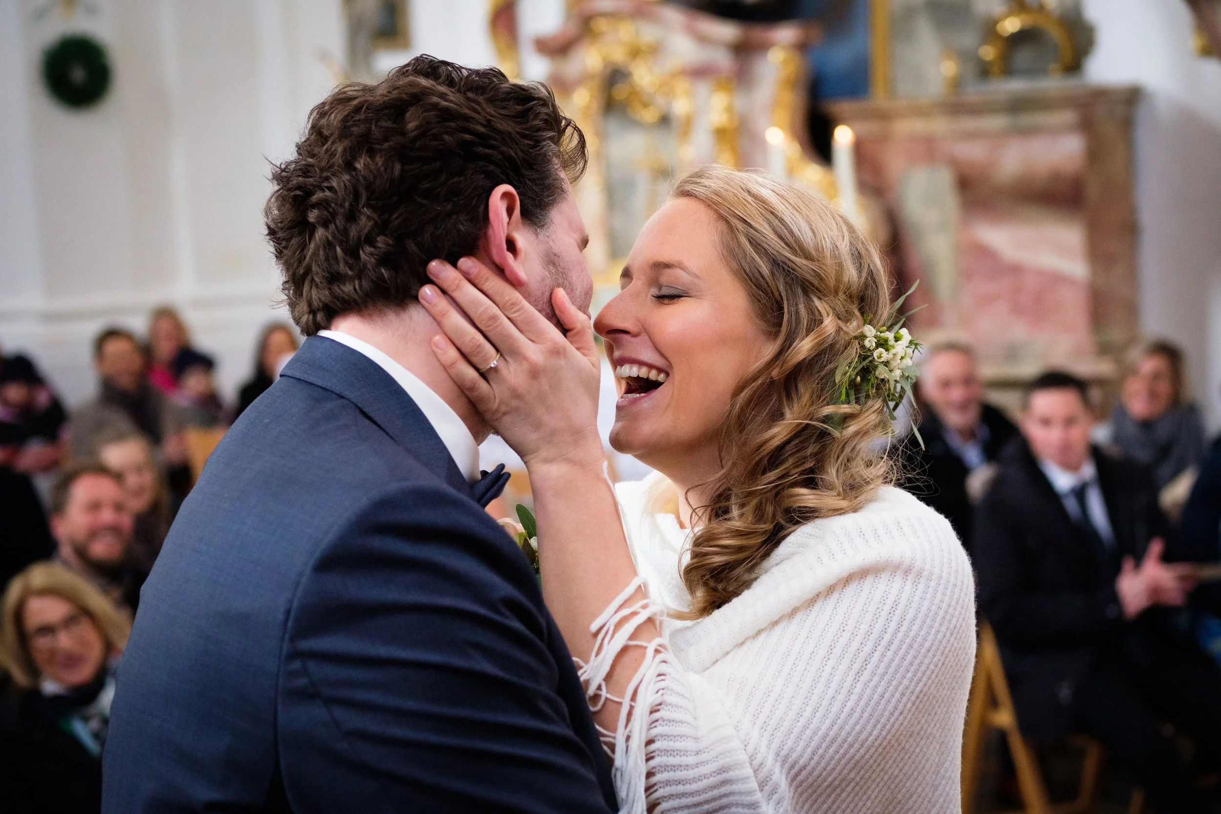 Ein Brautpaar bei einer Hochzeitszeremonie, das sich liebevoll berührt und umarmt, im Hintergrund sitzen Gäste in einer Kirche.