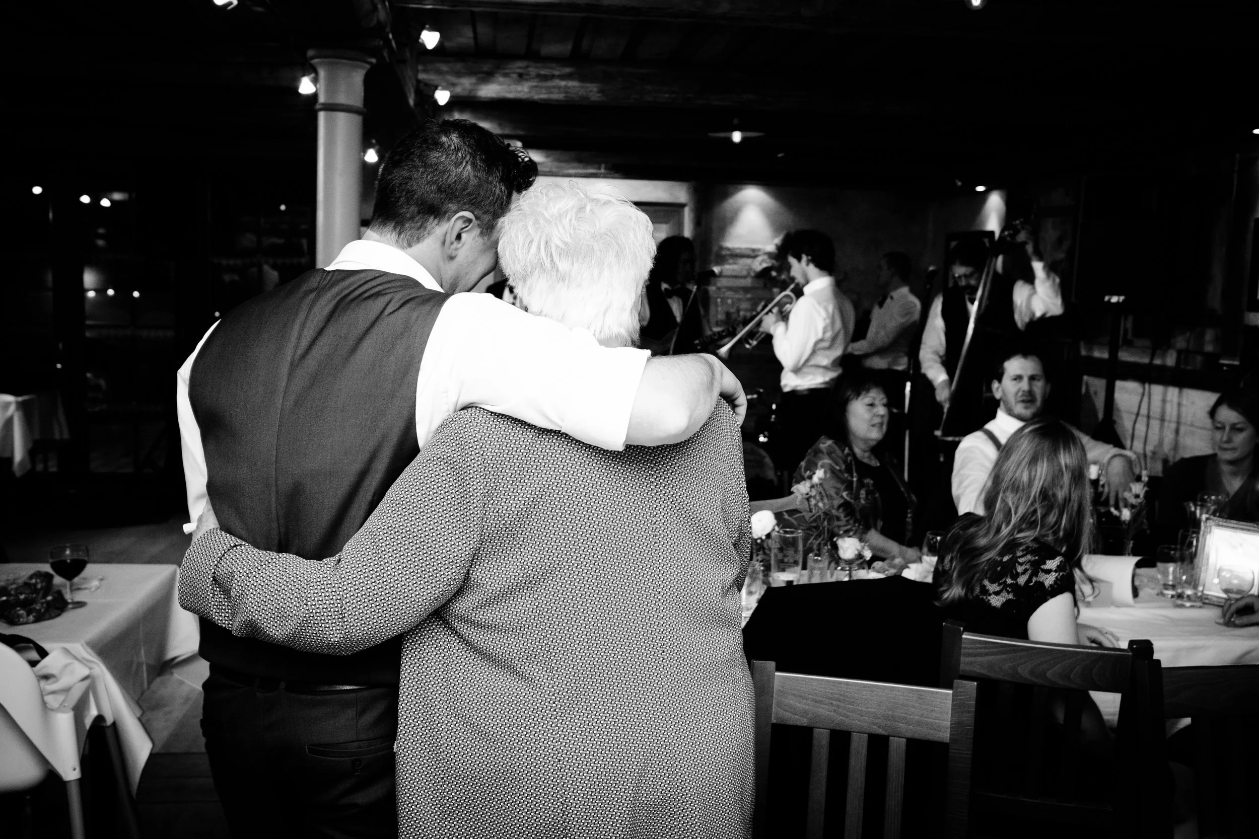 Zwei Menschen, ein jüngerer Mann und eine ältere Frau, umarmen sich bei einer Feier in einem Restaurant oder Saal, im Hintergrund eine Band mit Musikern.