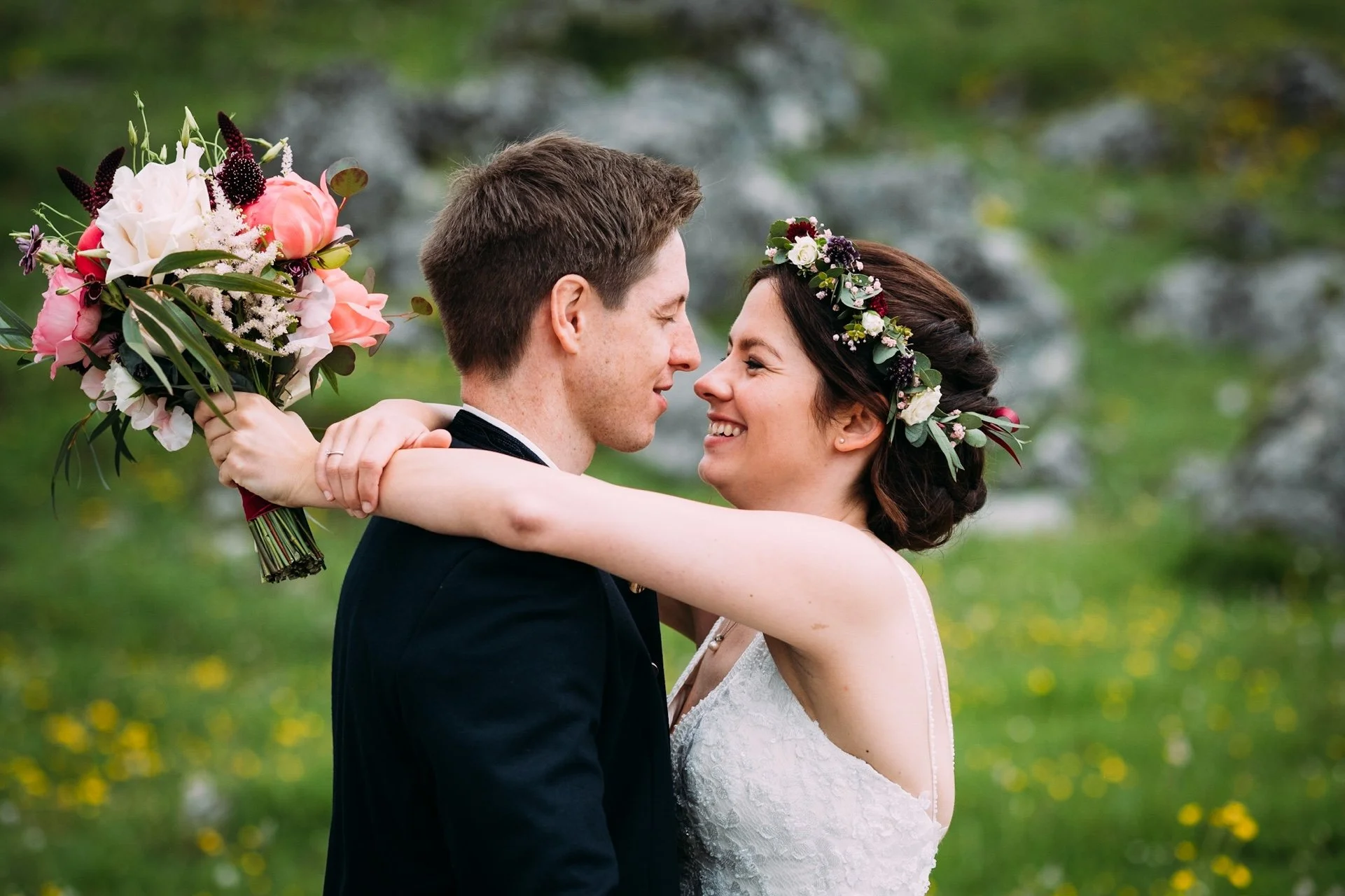 Ein Brautpaar umarmt sich im Grünen, die Braut trägt einen Blumenkranz und einen weißen Kleid, der Bräutigam trägt einen schwarzen Anzug.