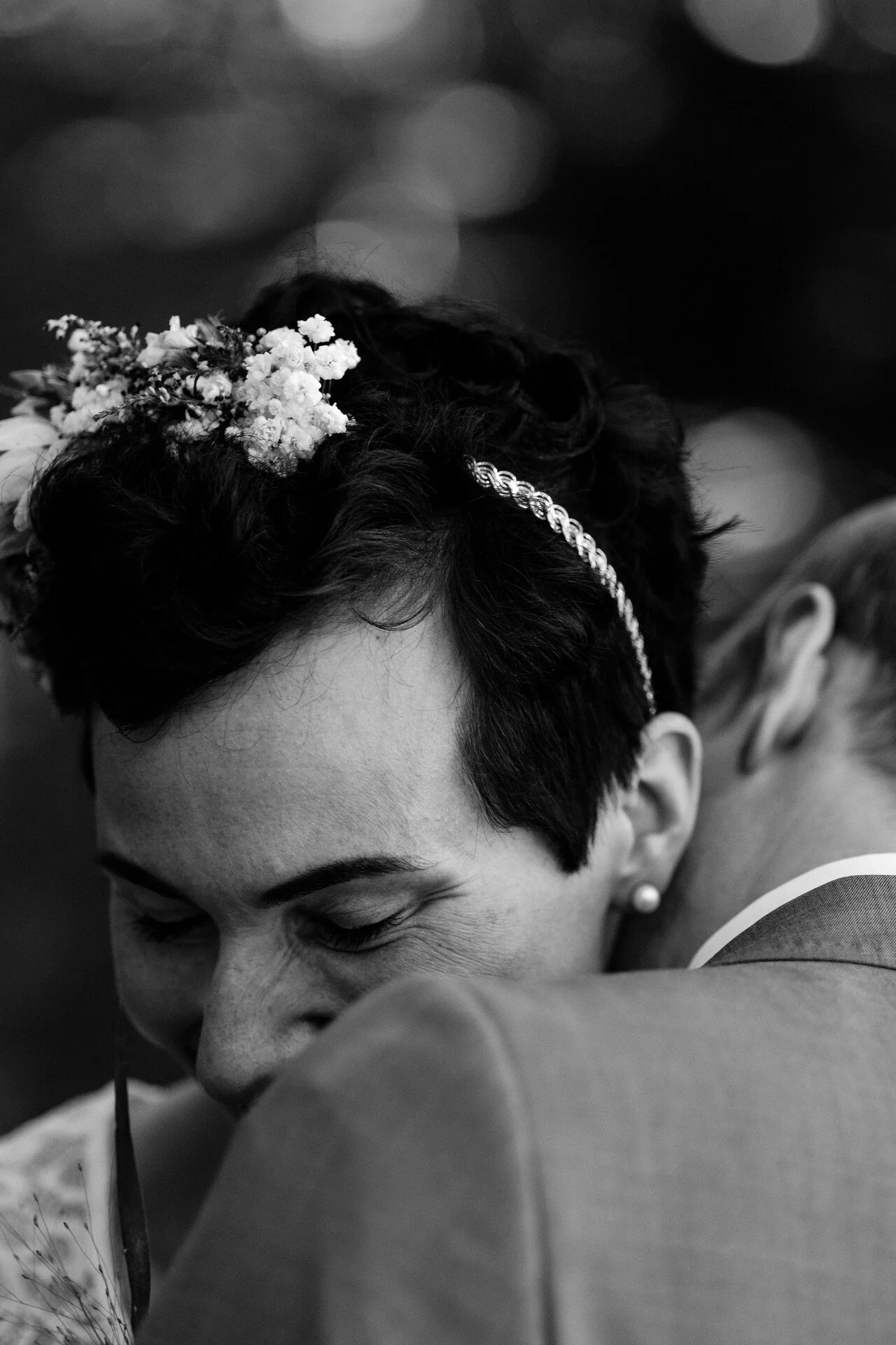 Eine schwarze und weiße Aufnahme eines glücklichen Menschen, der einen anderen umarmt, vermutlich bei einer Hochzeit, mit einem Blumenkranz im Haar und einem Kleid.