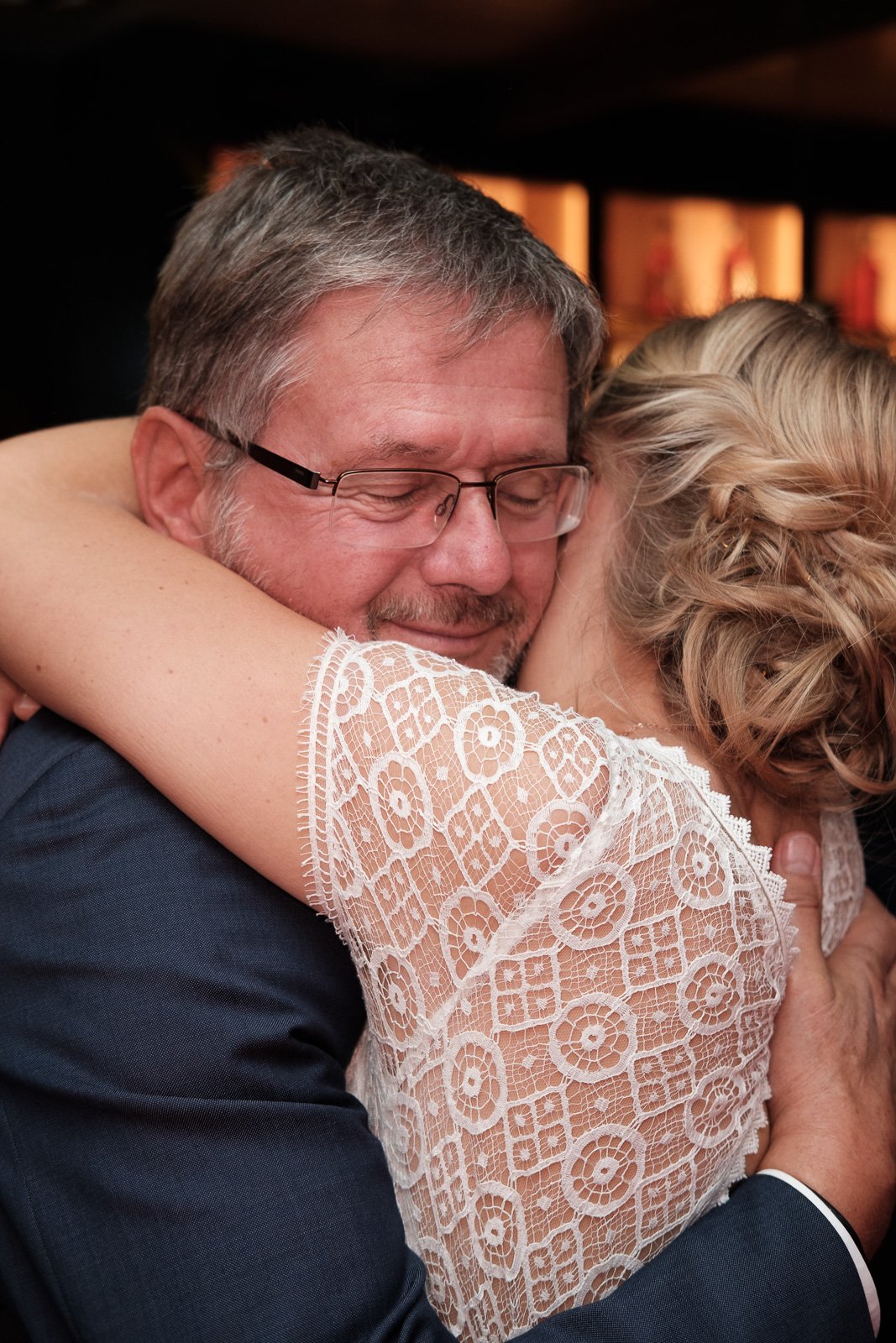 Ein Mann und eine Frau umarmen sich herzlich, das Gesicht des Mannes ist sichtbar, er hat graue Haare, trägt eine Brille und ein dunkles Sakko. Die Frau trägt ein weißes Spitzenkleid und hat blondes, lockiges Haar.