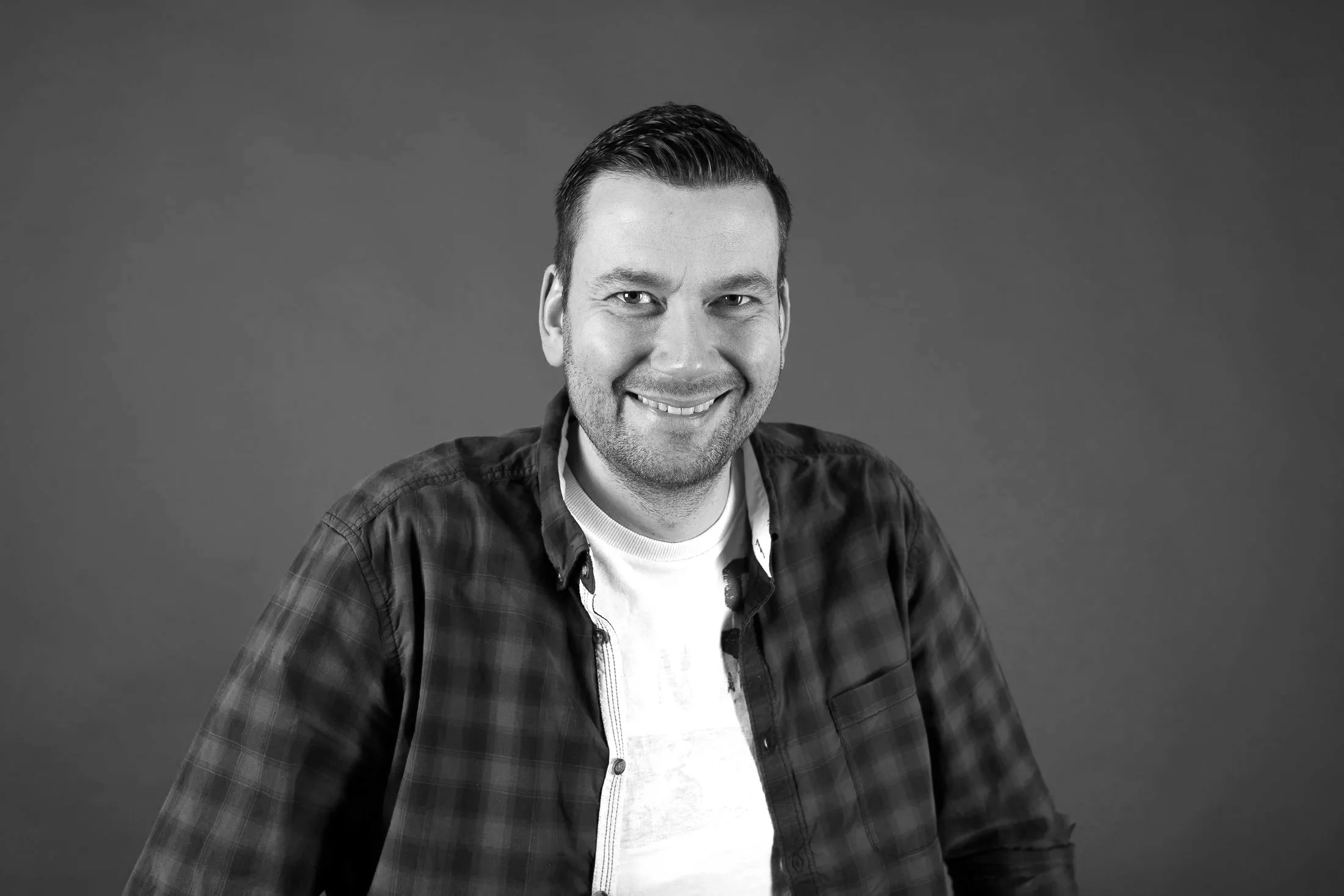 Schwarz-Weiß-Foto eines lächelnden Fotografen mit kurzen dunklen Haaren, Träger eines karierten Hemdes und weißem T-Shirt, vor einfachem grauem Hintergrund.