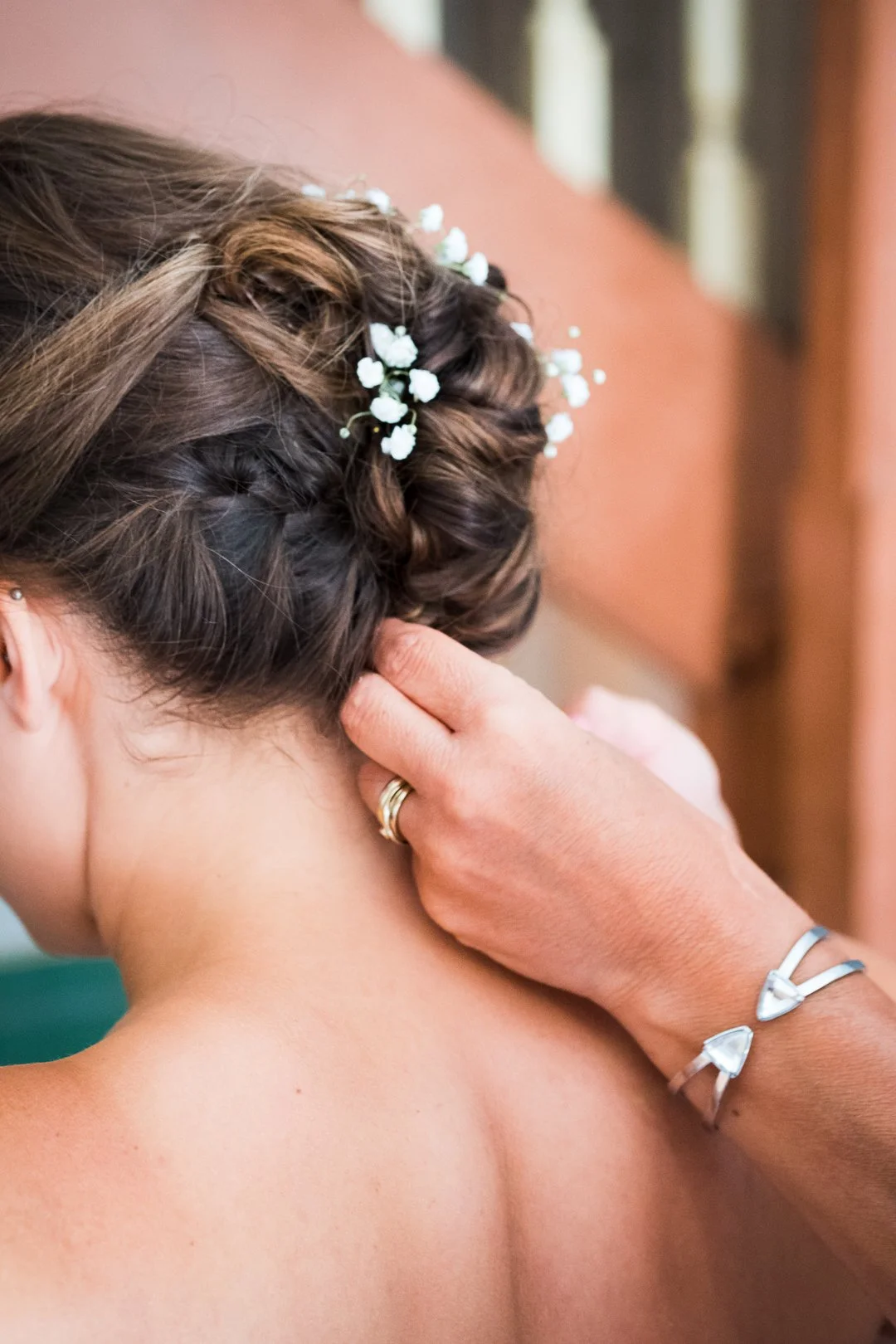 Eine Frau bekommt eine Hochsteckfrisur mit kleinen weißen Blumen am Hinterkopf, eine Hand mit einem Ring berührt den Nacken.