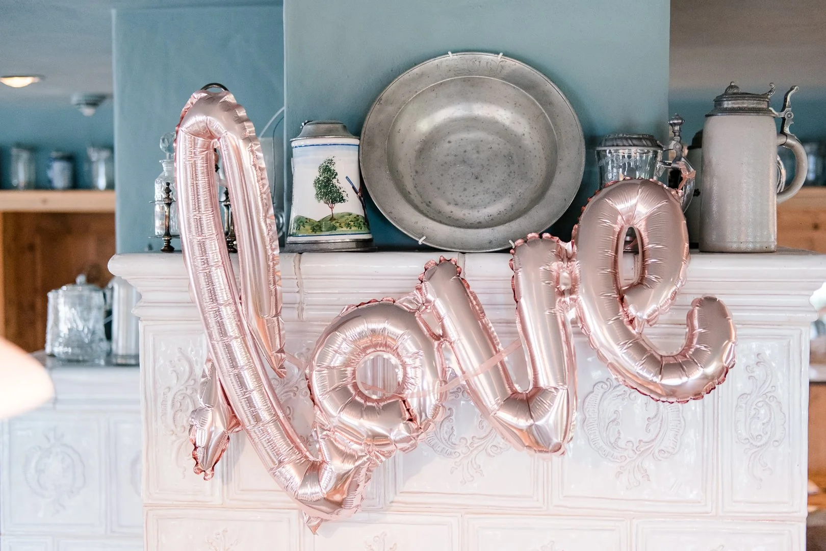 Dekoration mit rosafarbenen Ballonbuchstaben, die das Wort 'LOVE' bilden, vor einer weißen Kommode mit klassischen Schnitzereien. Auf der Kommode stehen alte Silber- und Keramikgefäße sowie eine große Metallplatte oberhalb. Der Hintergrund ist eine M