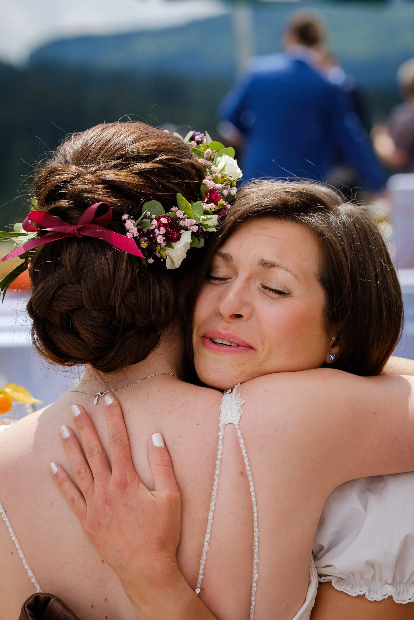 Zwei Frauen umarmen sich emotional. Eine trägt ein Brautkleid mit Spaghetti-Trägern und ein Blumengewinde im Haar, die andere hat kurze braune Haare und trägt elegante Ohrringe.
