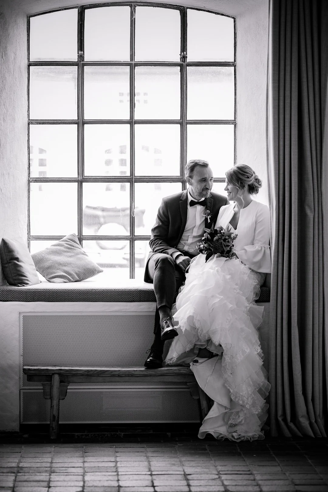 Braut und Bräutigam sitzen auf einer Fensterbank, schauen sich liebevoll an, sie hält einen Blumenstrauß, im Hintergrund eine große Fensterfront in einem Raum mit Vorhängen.
