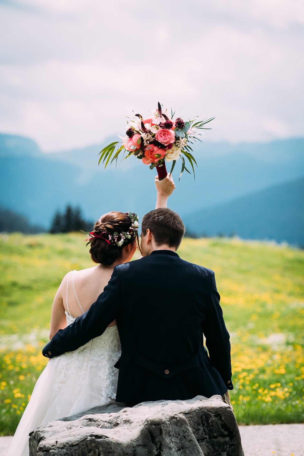 Ein Hochzeitsbild mit einem Brautpaar, das auf einem Felsen im Freien sitzt, den Rücken zeigt, die Braut trägt ein weißes Kleid und einen Blumenkranz, der Bräutigam trägt einen schwarzen Anzug, die Braut hält einen großen Blumenstrauß nach oben, im H