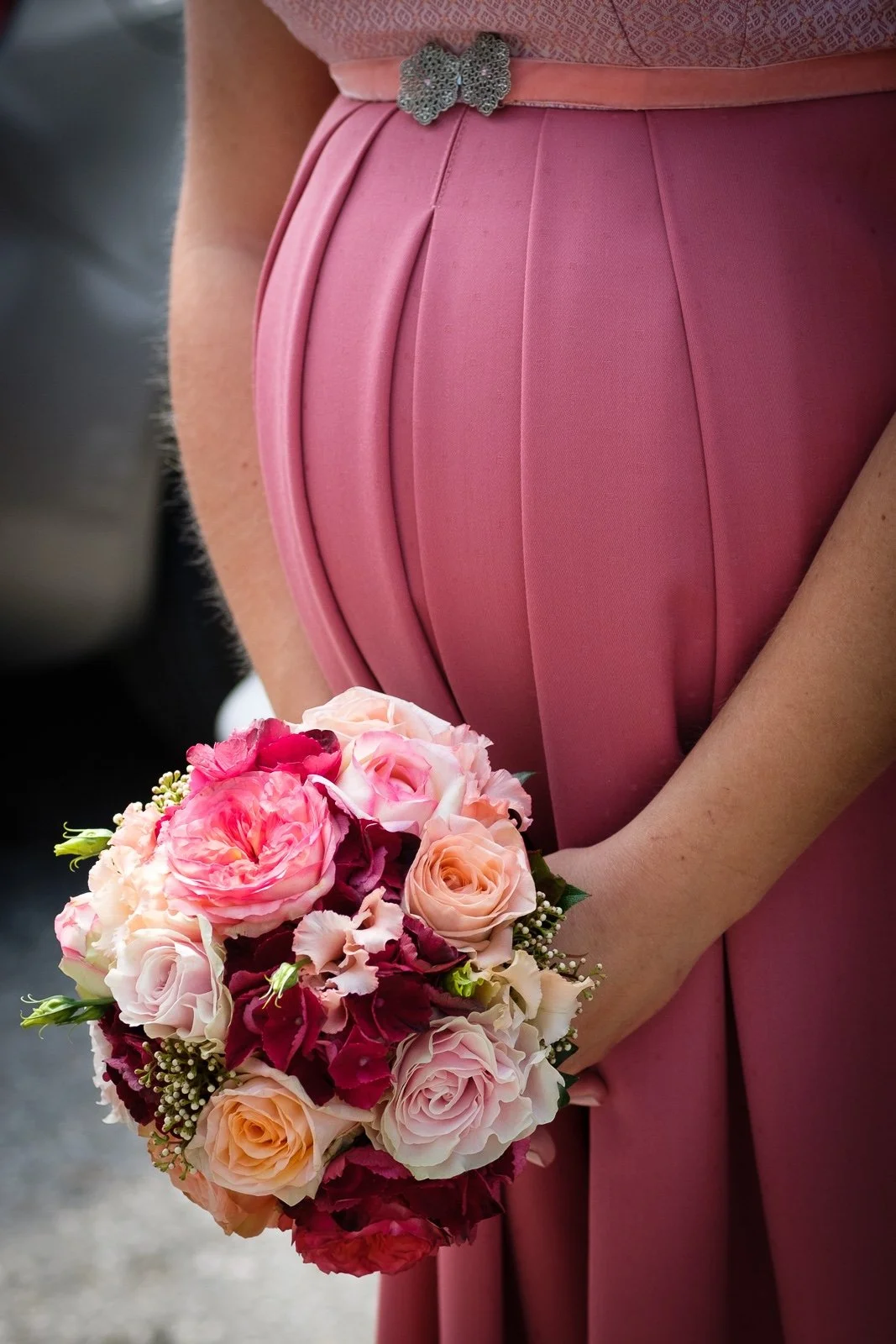 Nahaufnahme eines pinkfarbenen Kleides mit einem Blumen-Bross und einer Hand, die einen bunten Blumenstrauß hält.