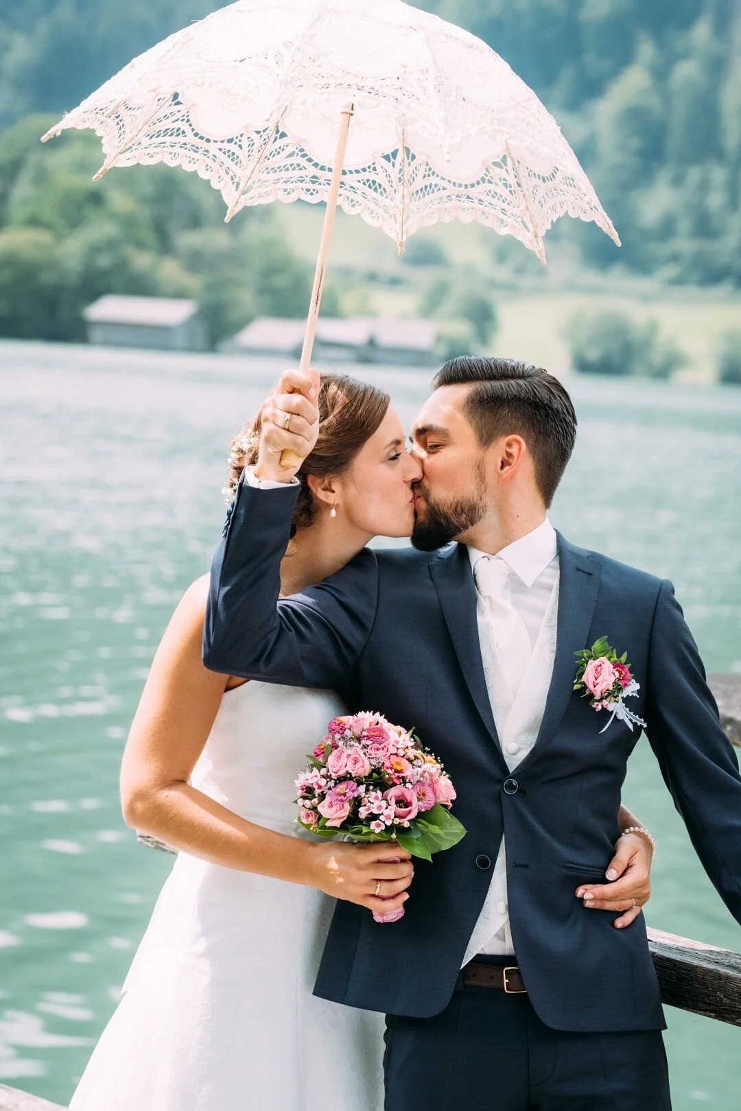Ein Brautpaar küsst sich am Wasser, die Braut hält einen Blumenstrauß aus rosa Blüten und bunten Blumen, während der Bräutigam einen weißen Kuss erhält. Die Braut trägt ein weißes Kleid, der Bräutigam einen dunklen Anzug, und sie halten einen weißen 