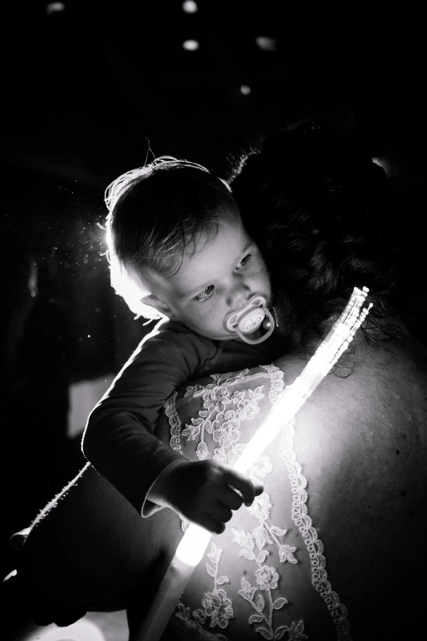 Ein kleines Kind mit Schnuller liegt auf einer Schulter, hält ein leuchtendes Lichtstäbchen und sieht nach unten am Hochzeitskleid vorbei.