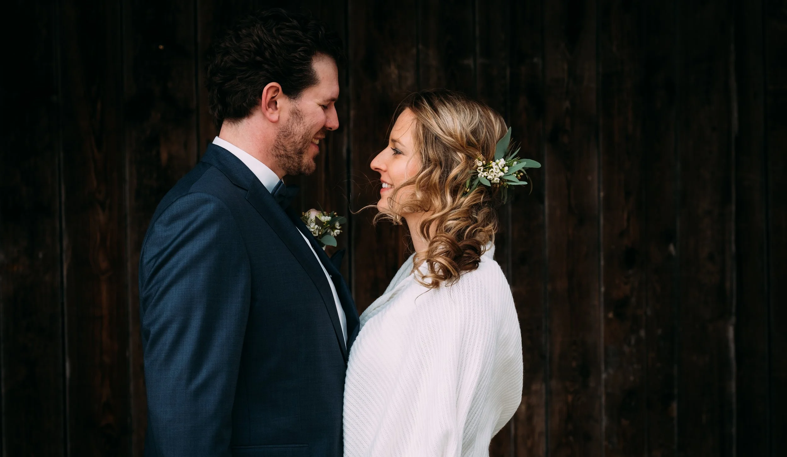Ein Mann und eine Frau stehen sich bei einer Hochzeit gegenüber, lächeln sich an, vor einem dunklen Holz-Hintergrund.