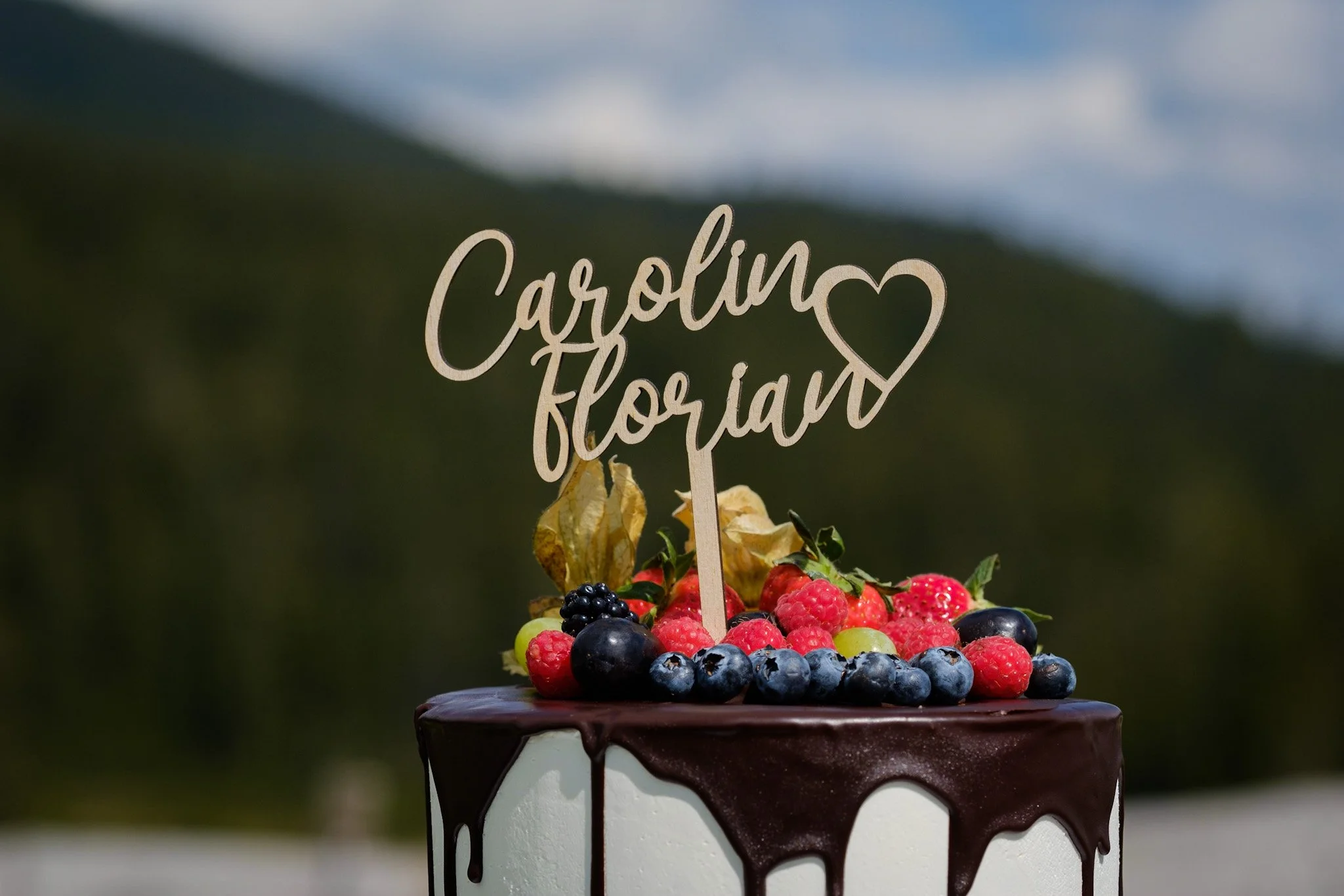 Ein festlicher Kuchen mit dunkler Schokoladenglasur, garniert mit bunten Beeren und einem Holzname 'Carolina Florian' als Dekoration.