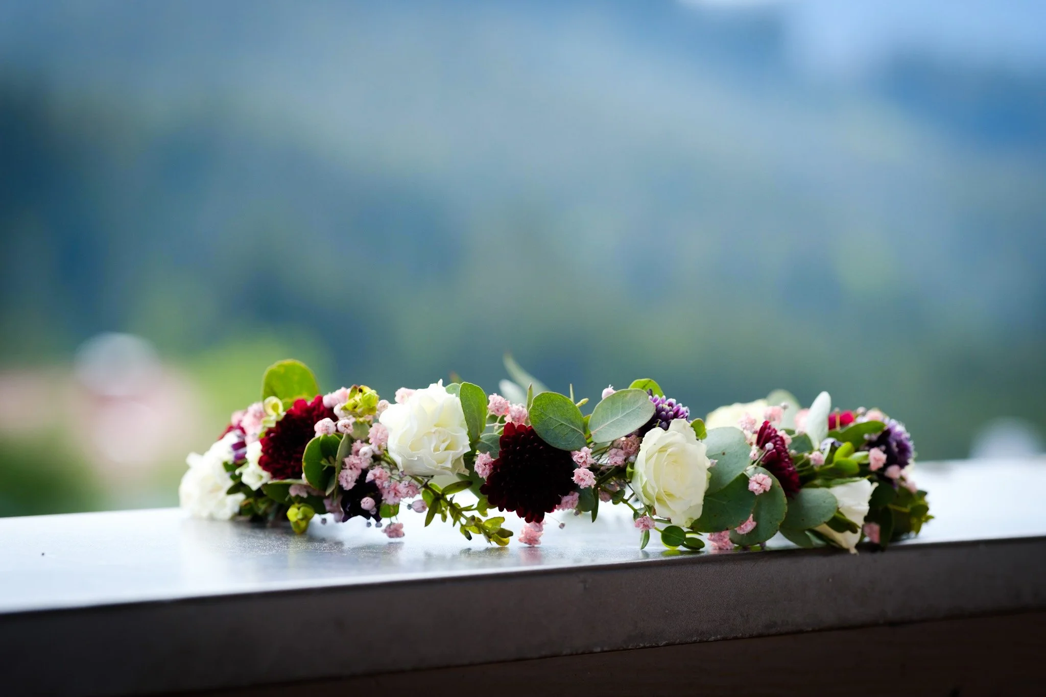 Blumenkranz mit weißen, dunkelroten und pinken Blumen liegt auf einer schwarzen Oberfläche, unscharfer Hintergrund mit bläulichem Himmel