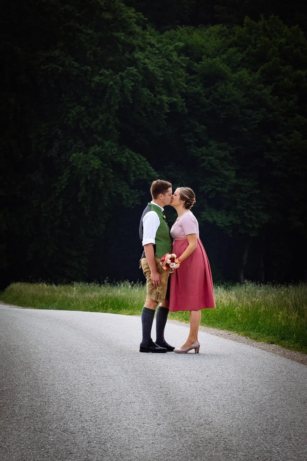 Ein junges Paar beim Kuss auf einer Landstraße, der Mann trägt traditionelle bayerische Kleidung und die Frau ein rotes Dirndl, sie hält einen Blumenstrauß.