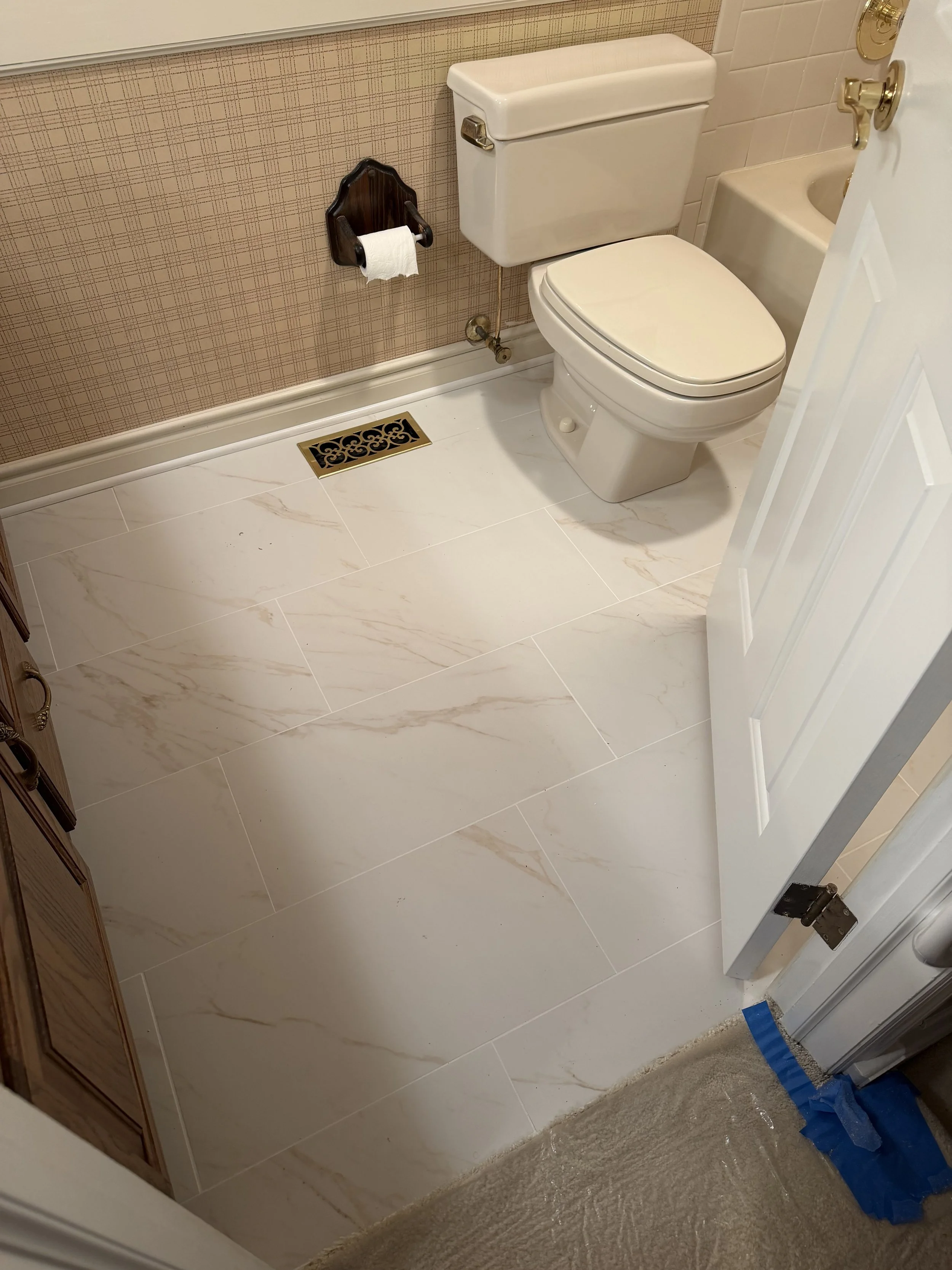 A small bathroom with beige patterned wallpaper, a white toilet, a wooden toilet paper holder with a roll, a vent on the floor, and part of a white door open to the room.