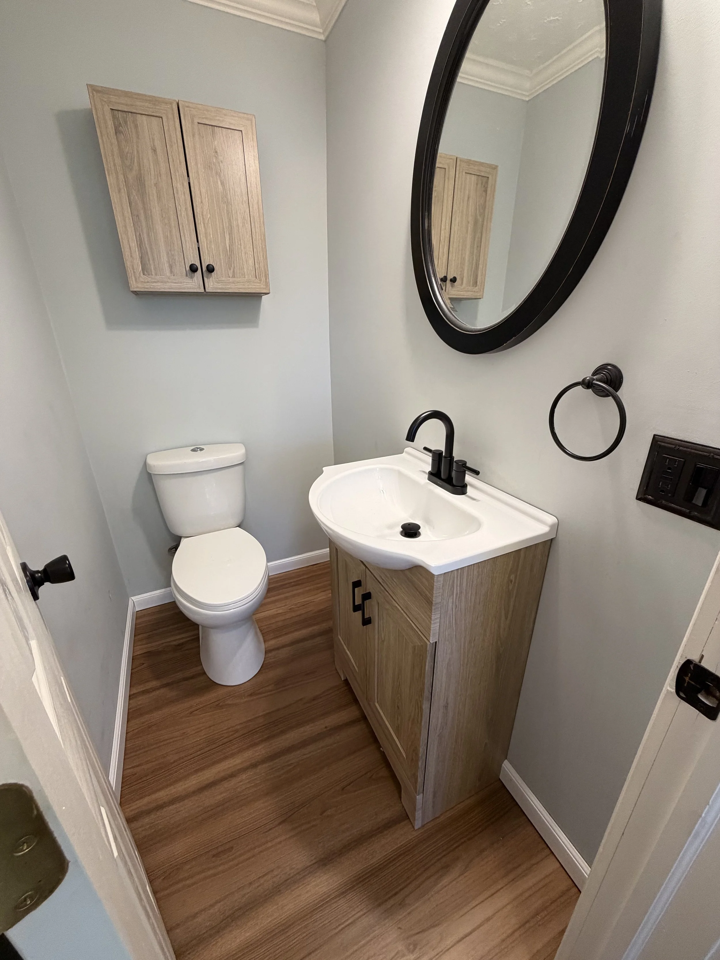 Small bathroom with a white toilet, a wooden vanity with a white sink, an oval mirror, a wooden cabinet above the toilet, a black faucet and towel ring, and wood-style flooring.